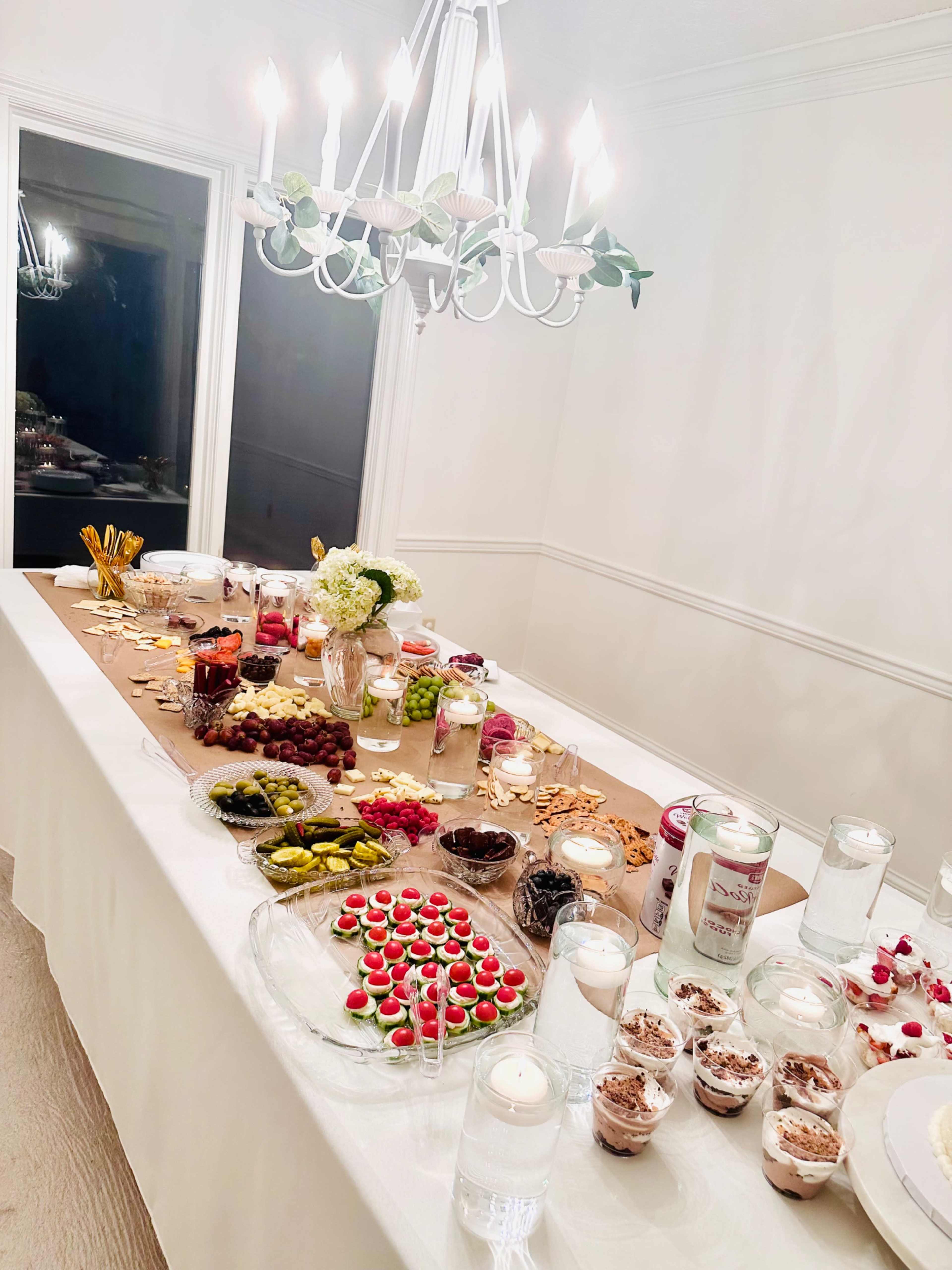 The image shows a dining table set with various foods, including fruits, desserts, and snacks, beneath a chandelier.