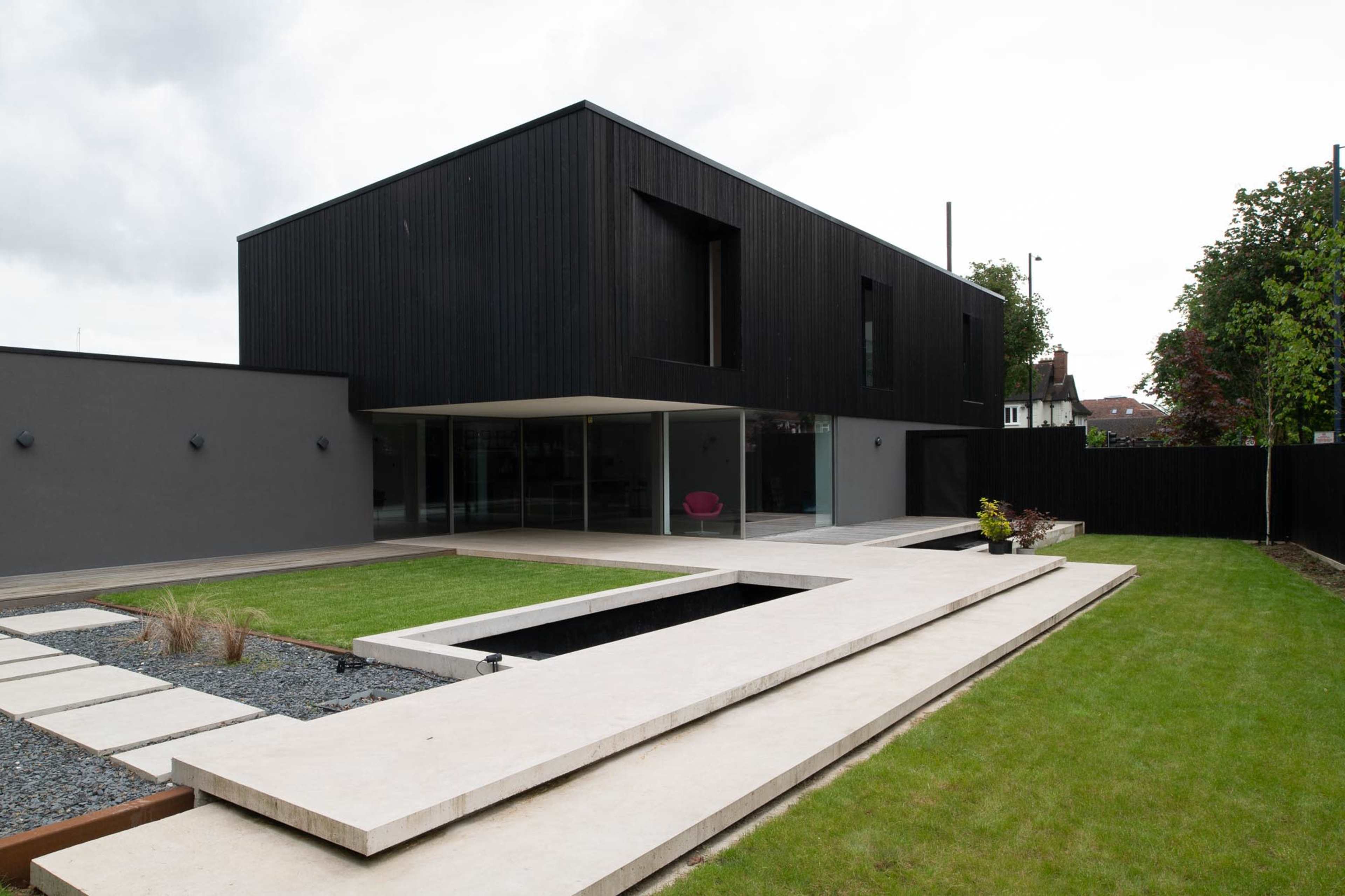 A modern black wooden house with large glass windows is surrounded by a landscaped yard featuring a rectangular stone path and a small water feature.