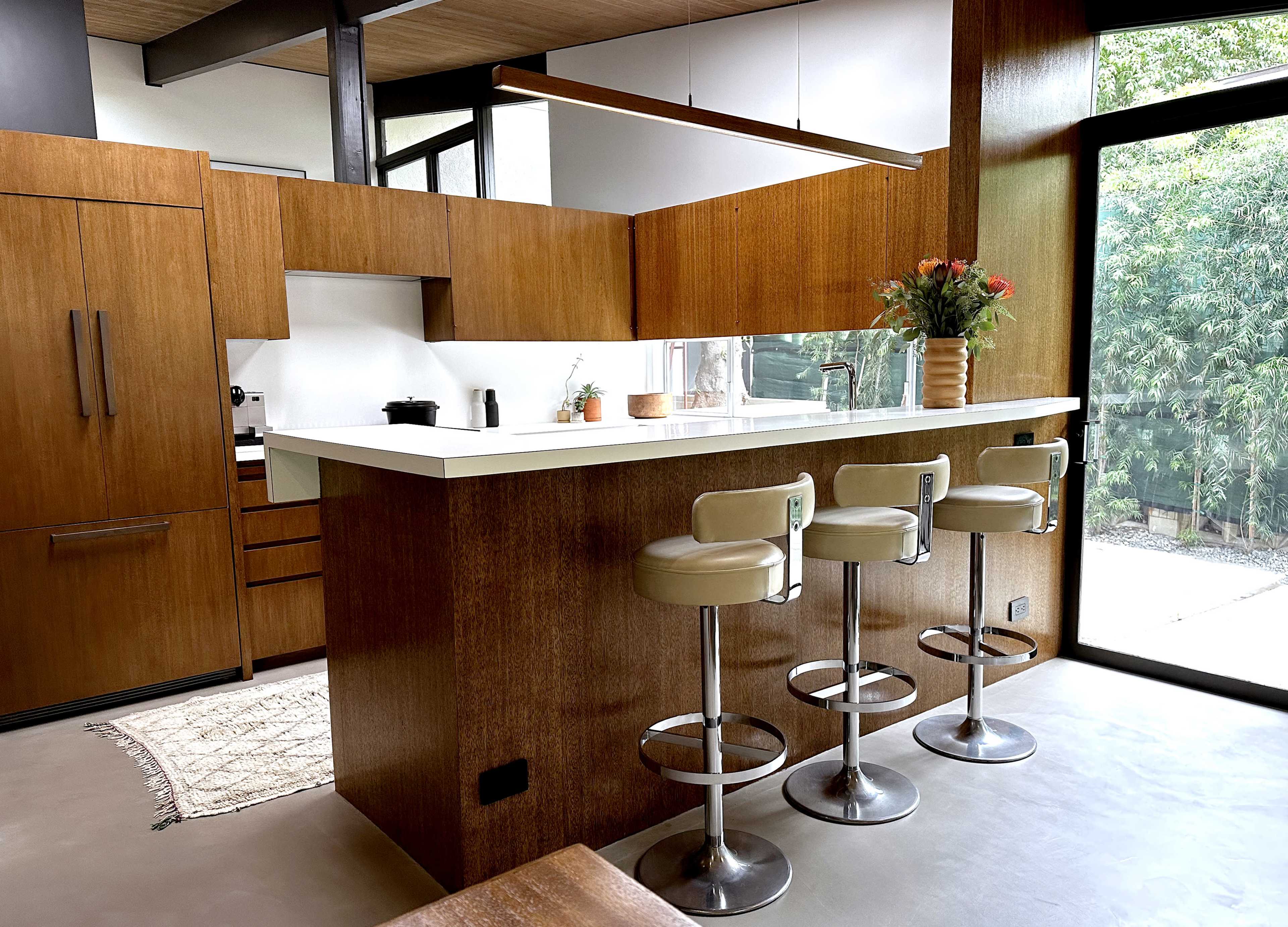 The image shows a modern kitchen featuring wooden cabinetry, a white countertop, and three bar stools at a kitchen island.