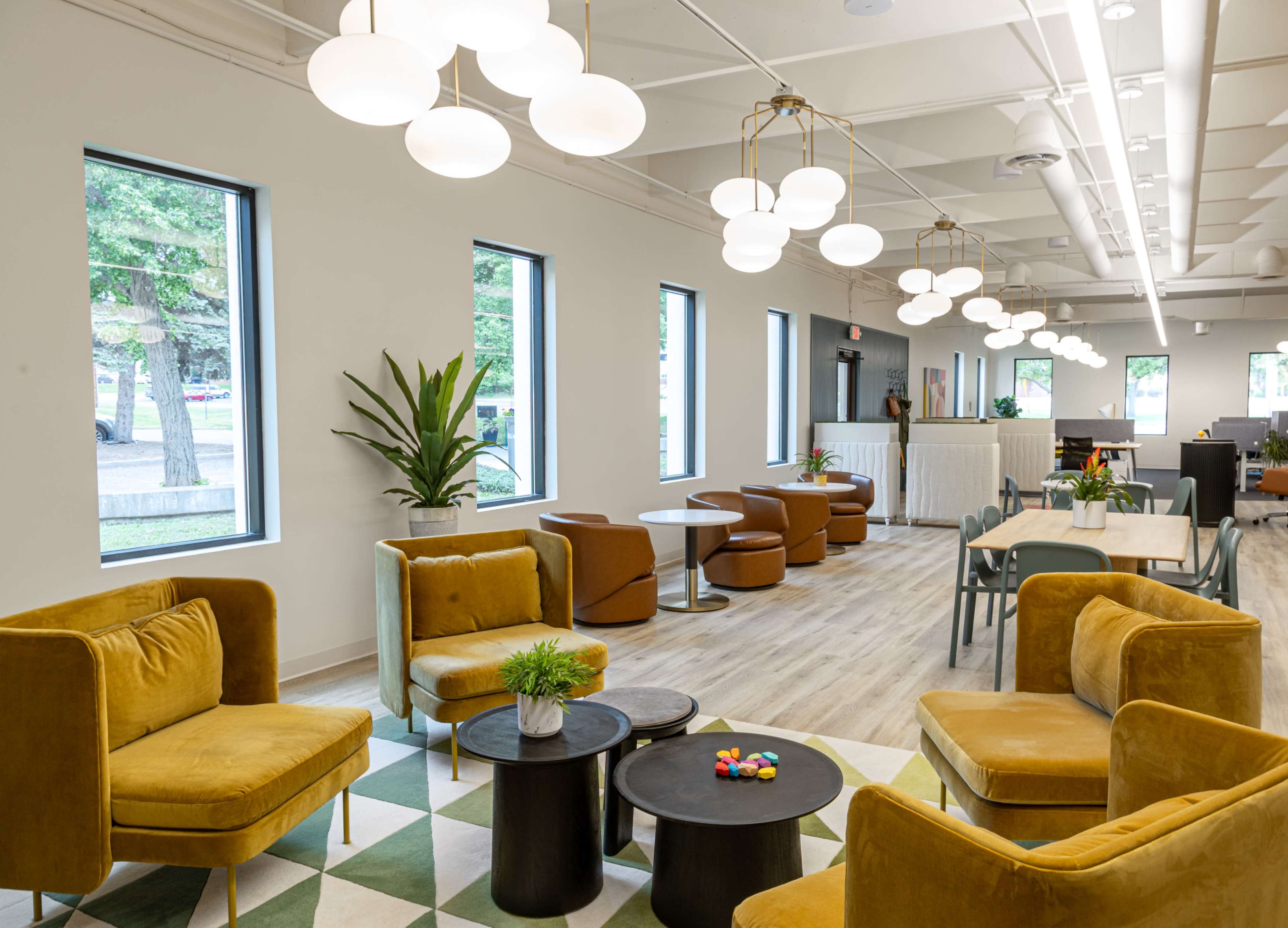 The image shows a modern lounge area featuring yellow and brown seating, round tables, and overhead lighting, with large windows allowing natural light to enter.