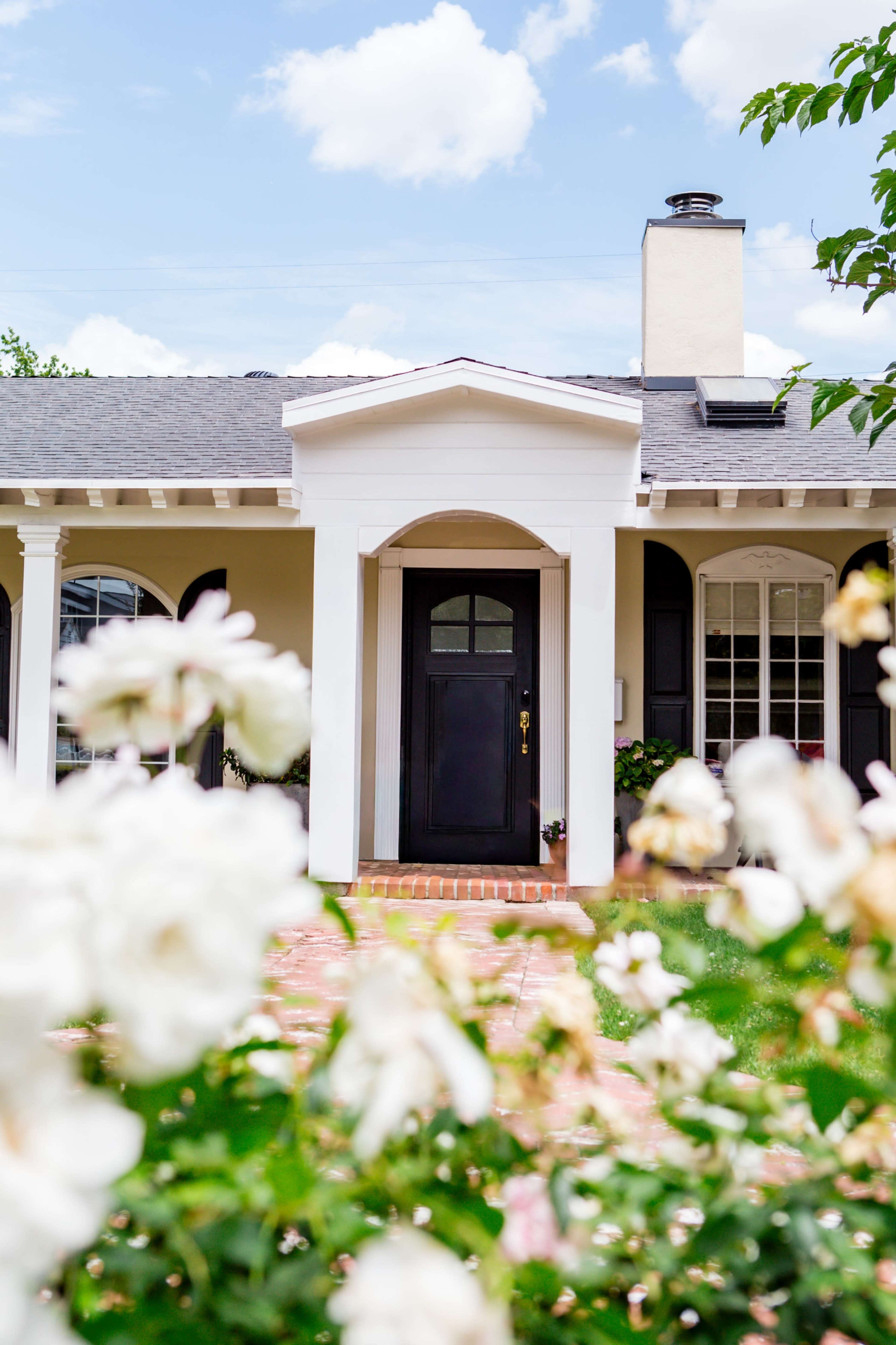 The image shows a house with a black front door framed by white columns, surrounded by blooming white roses and a clear blue sky.