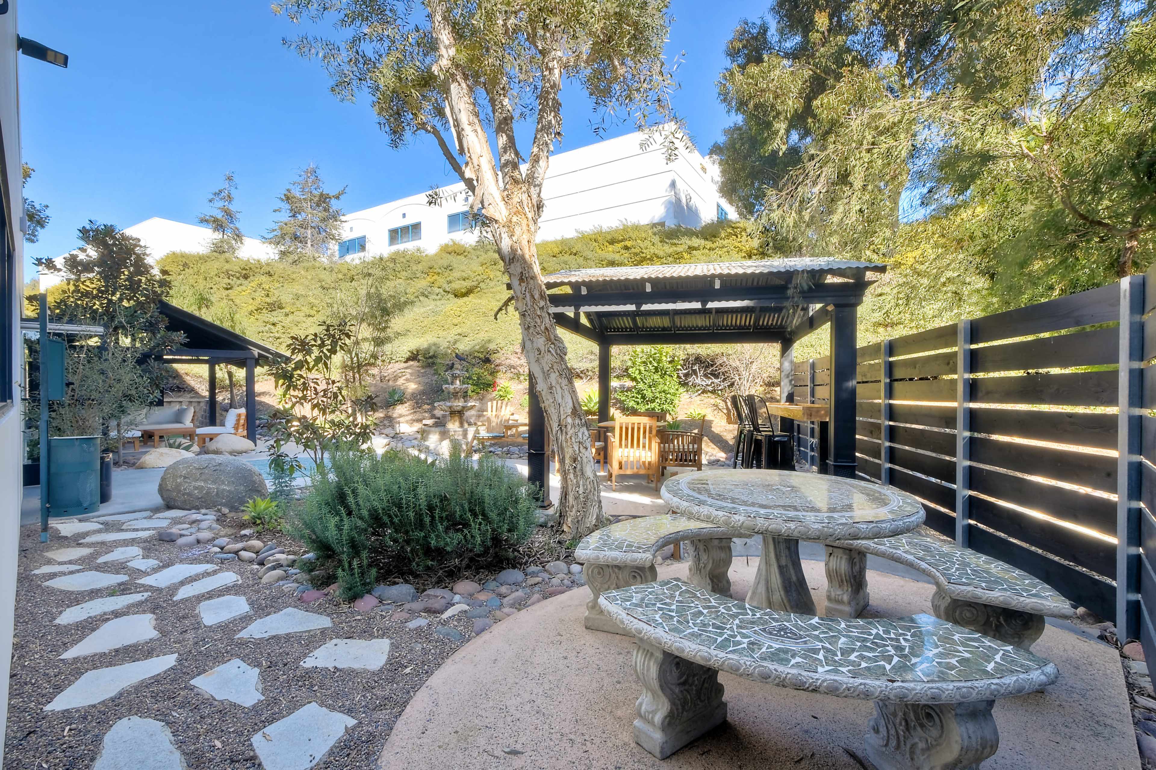 The image shows a landscaped outdoor area featuring a stone table with bench seating, a gazebo, and garden vegetation.