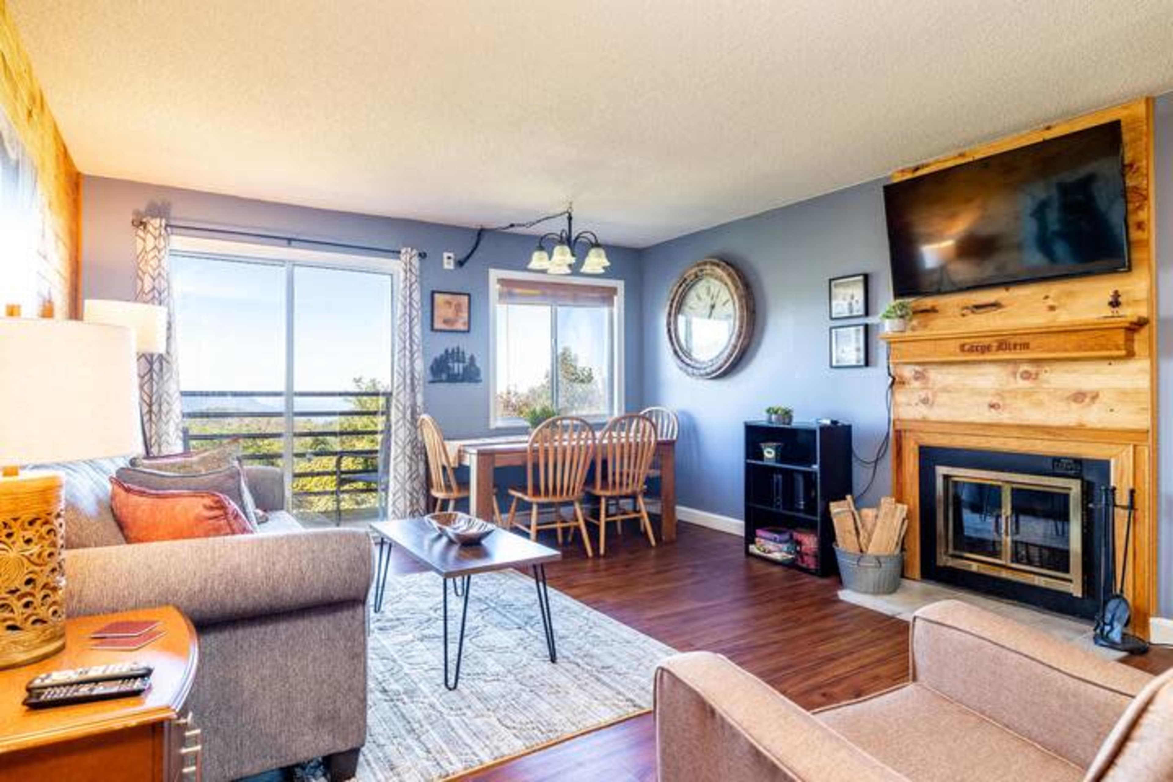 The image shows a modern living room with a cozy seating area, a dining table, and a fireplace, all accented by large windows letting in natural light.