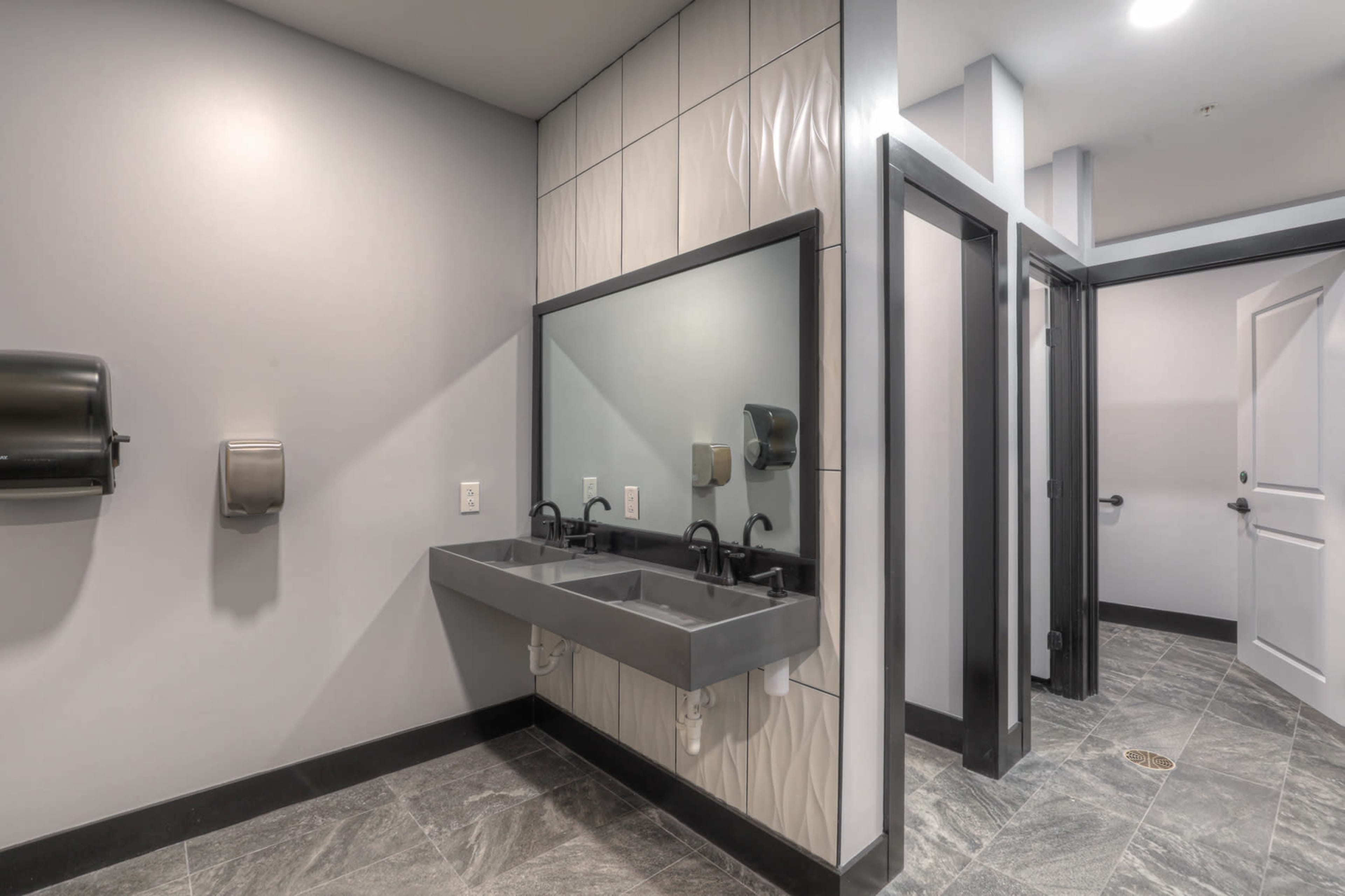The image shows a modern restroom with a double sink, a large mirror, and adjacent restroom stalls.