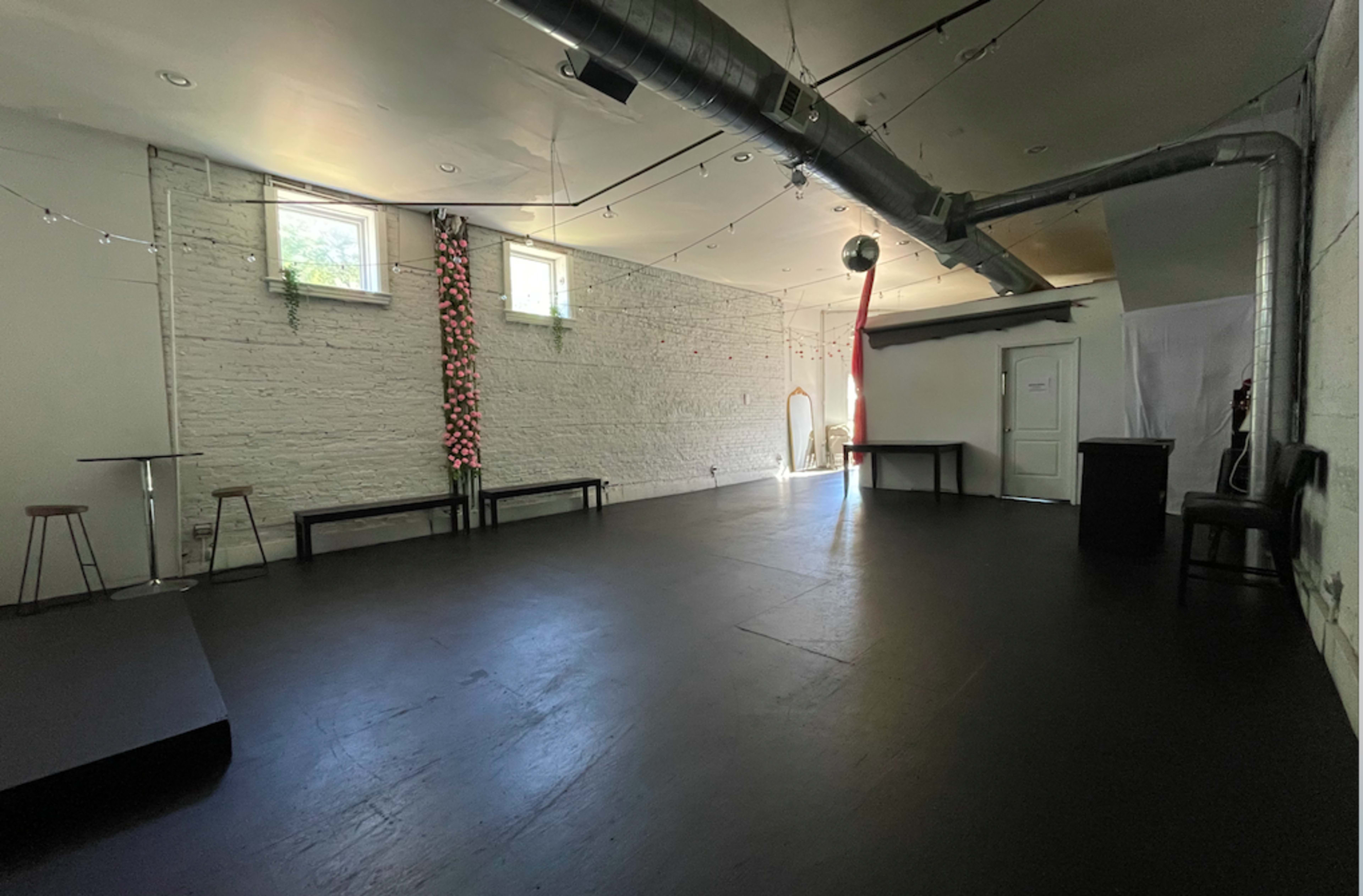 The image shows a spacious, empty room with a dark floor, white brick walls, and several tables along the perimeter, illuminated by natural light from windows.