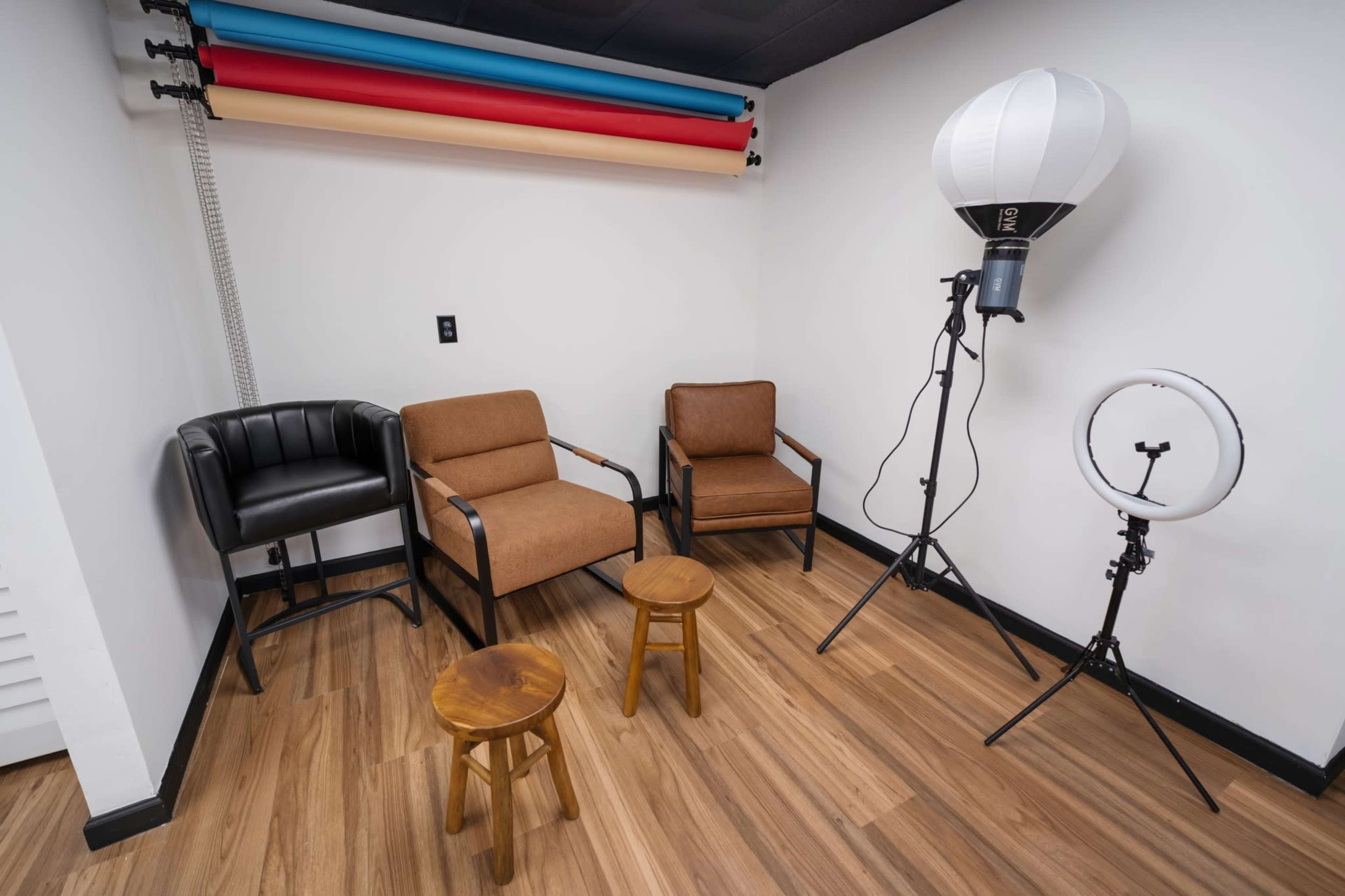 The image shows a small photo studio with two chairs, three stools, and lighting equipment against a white wall.
