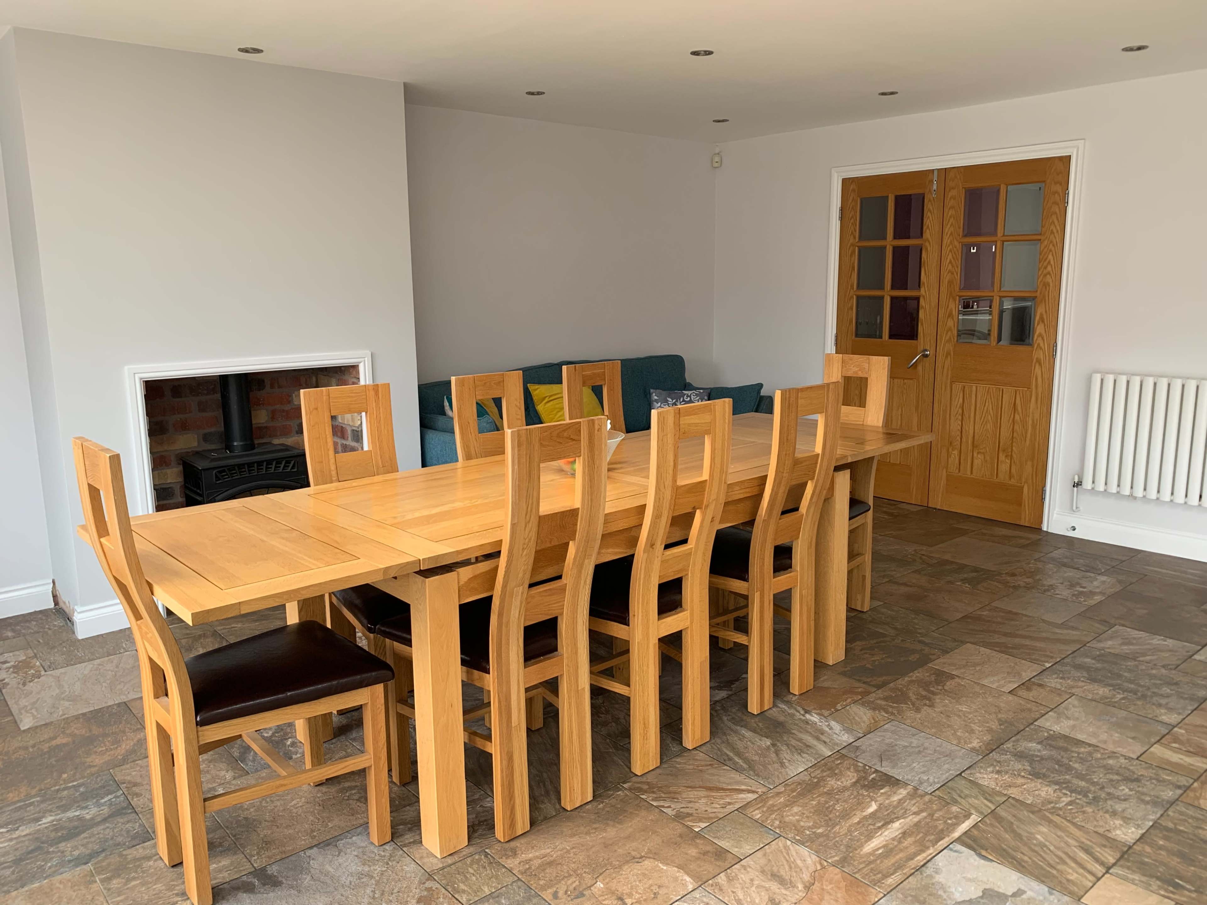 The image shows a dining area featuring a large wooden table surrounded by matching chairs, with a sofa and door in the background.