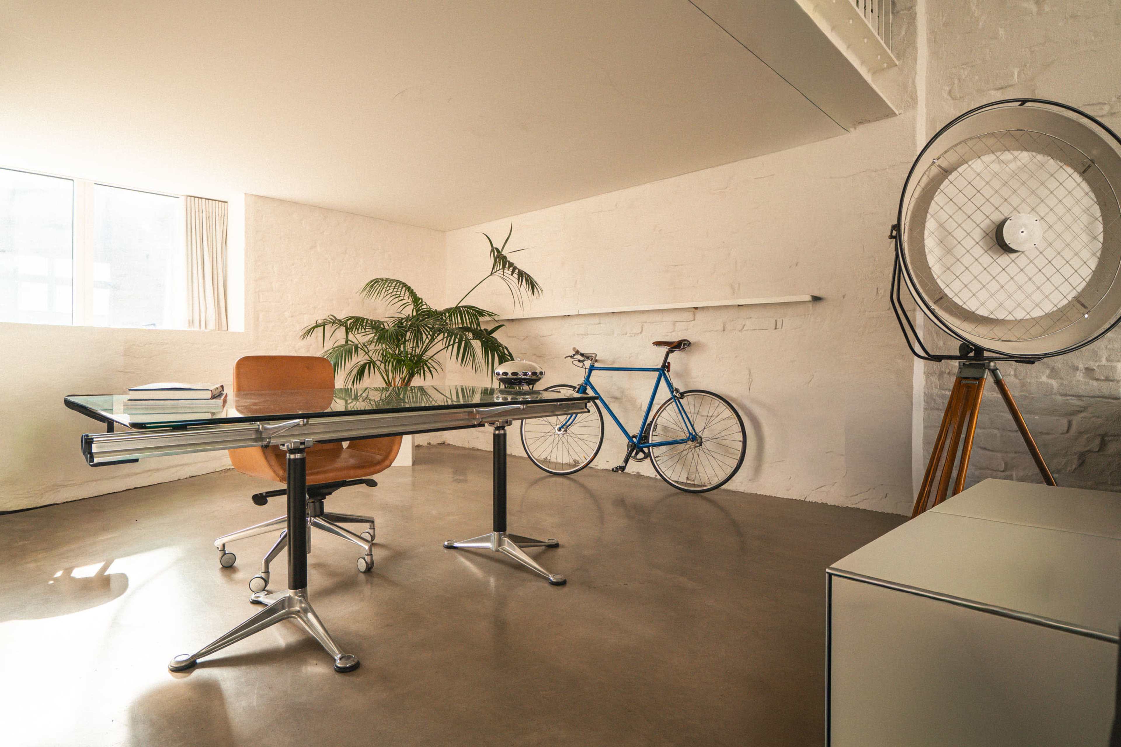 A modern office space features a glass desk, an ergonomic chair, a bicycle against the wall, and a large fan beside a potted plant.
