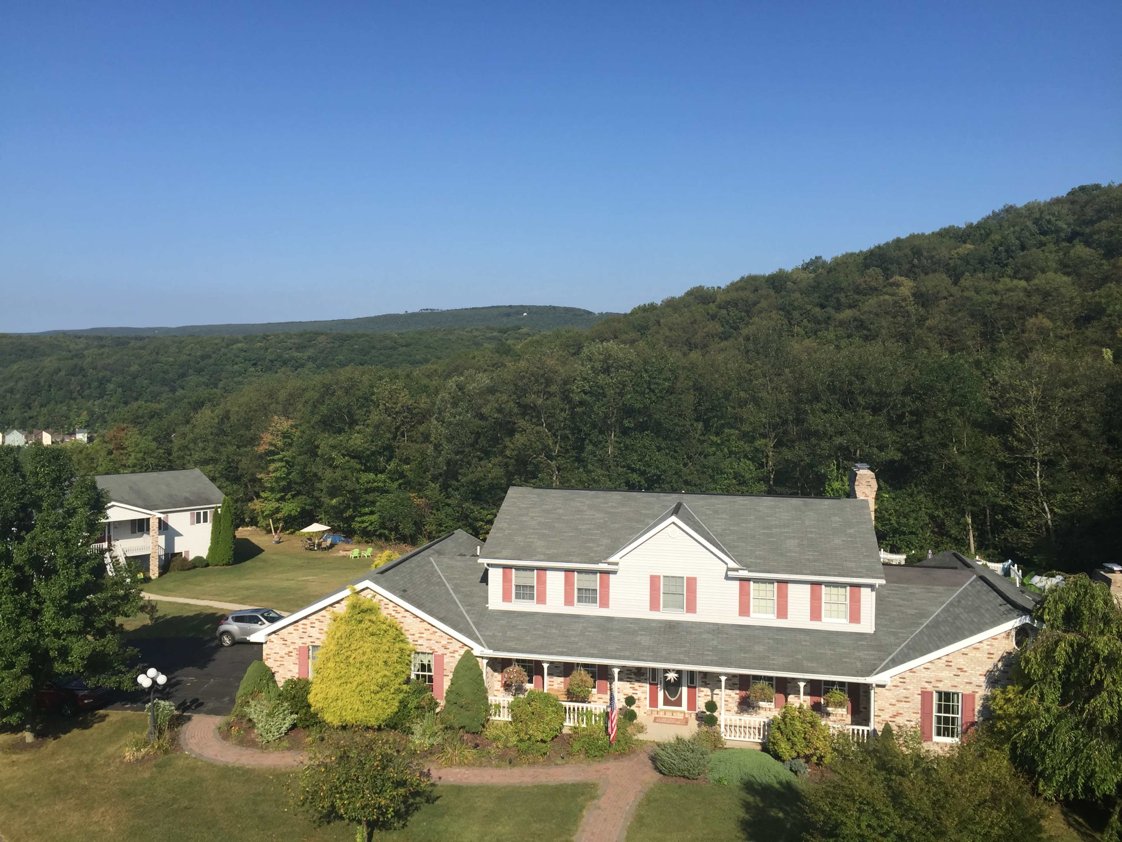 A two-story brick house with a gabled roof, surrounded by greenery and hills under a clear blue sky.