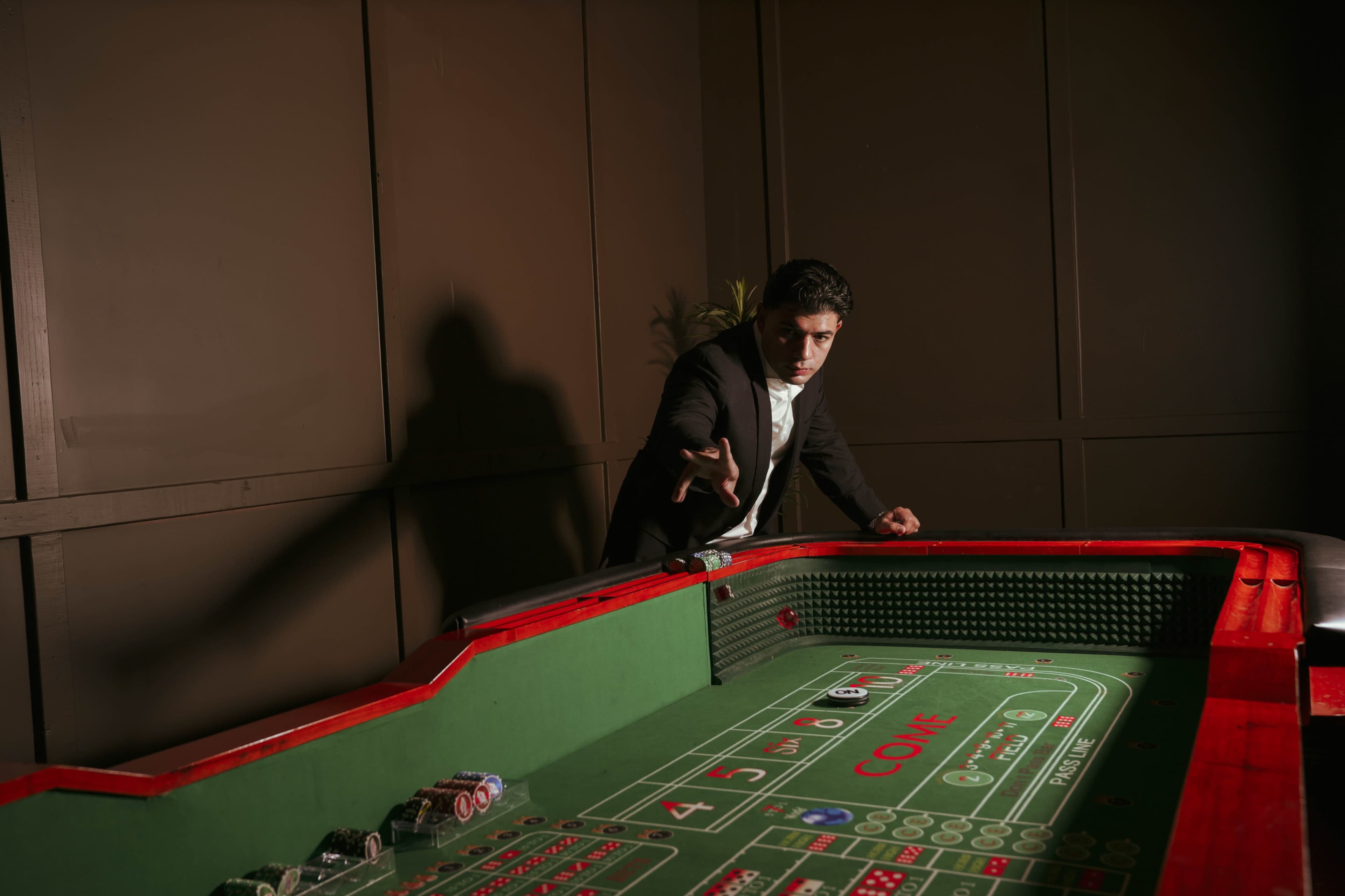 A man in a suit leans over a green craps table, gesturing as he prepares to roll dice.