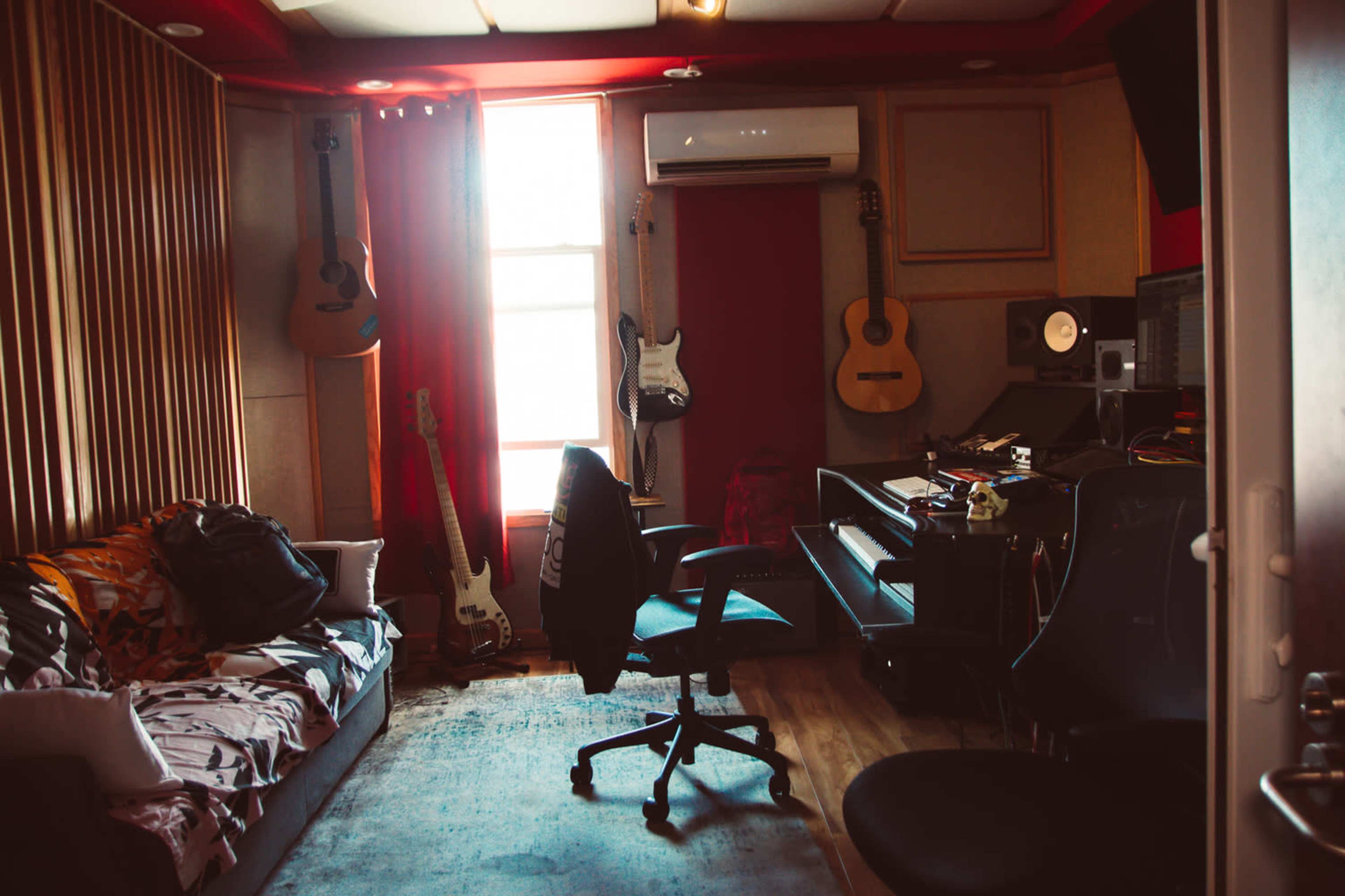 A music studio features a couch, various guitars on the walls, a piano, and recording equipment, with large windows providing natural light.