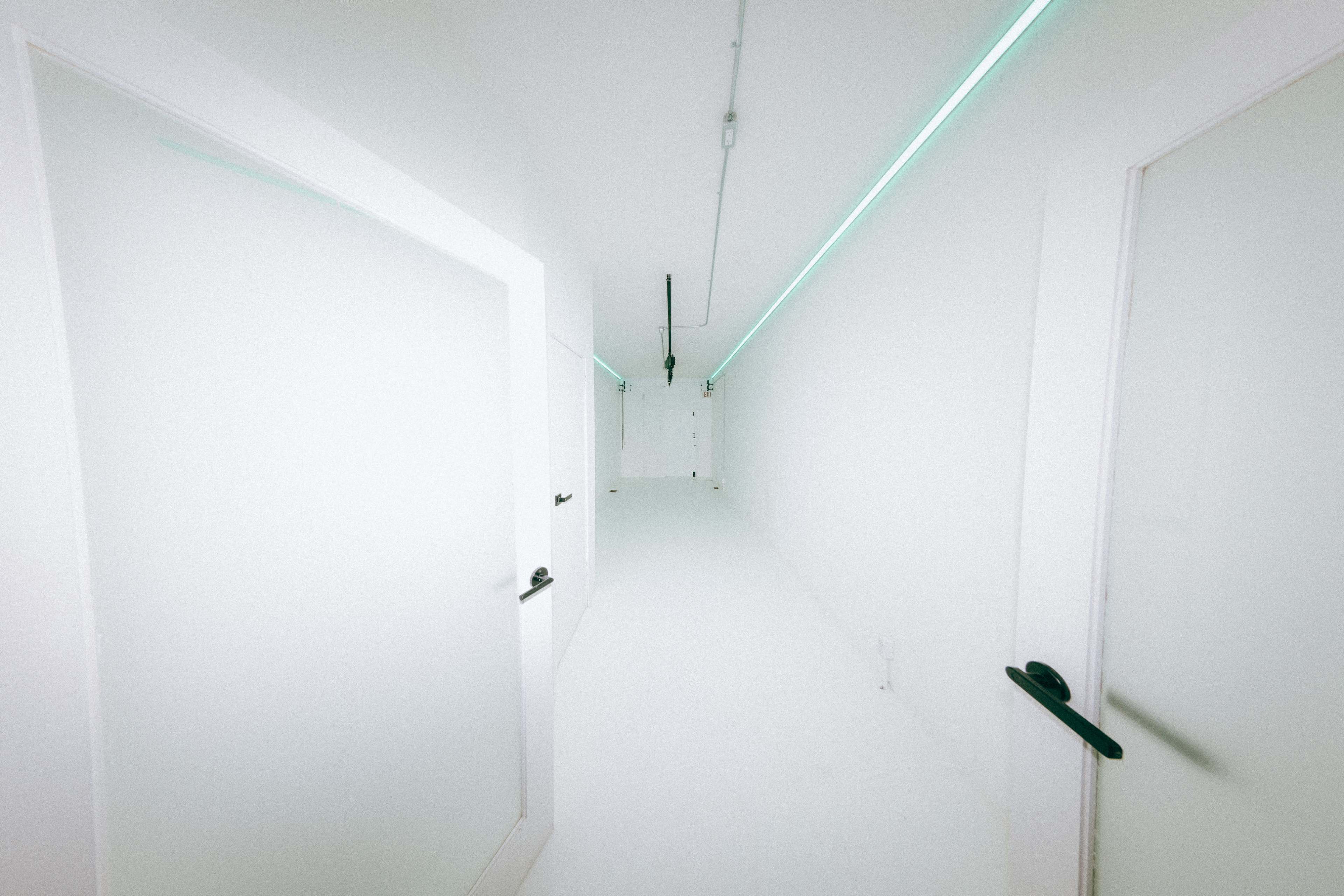 A long, narrow corridor with white walls and floors, featuring several closed doors and illuminated by a strip of blue-green light along the ceiling.
