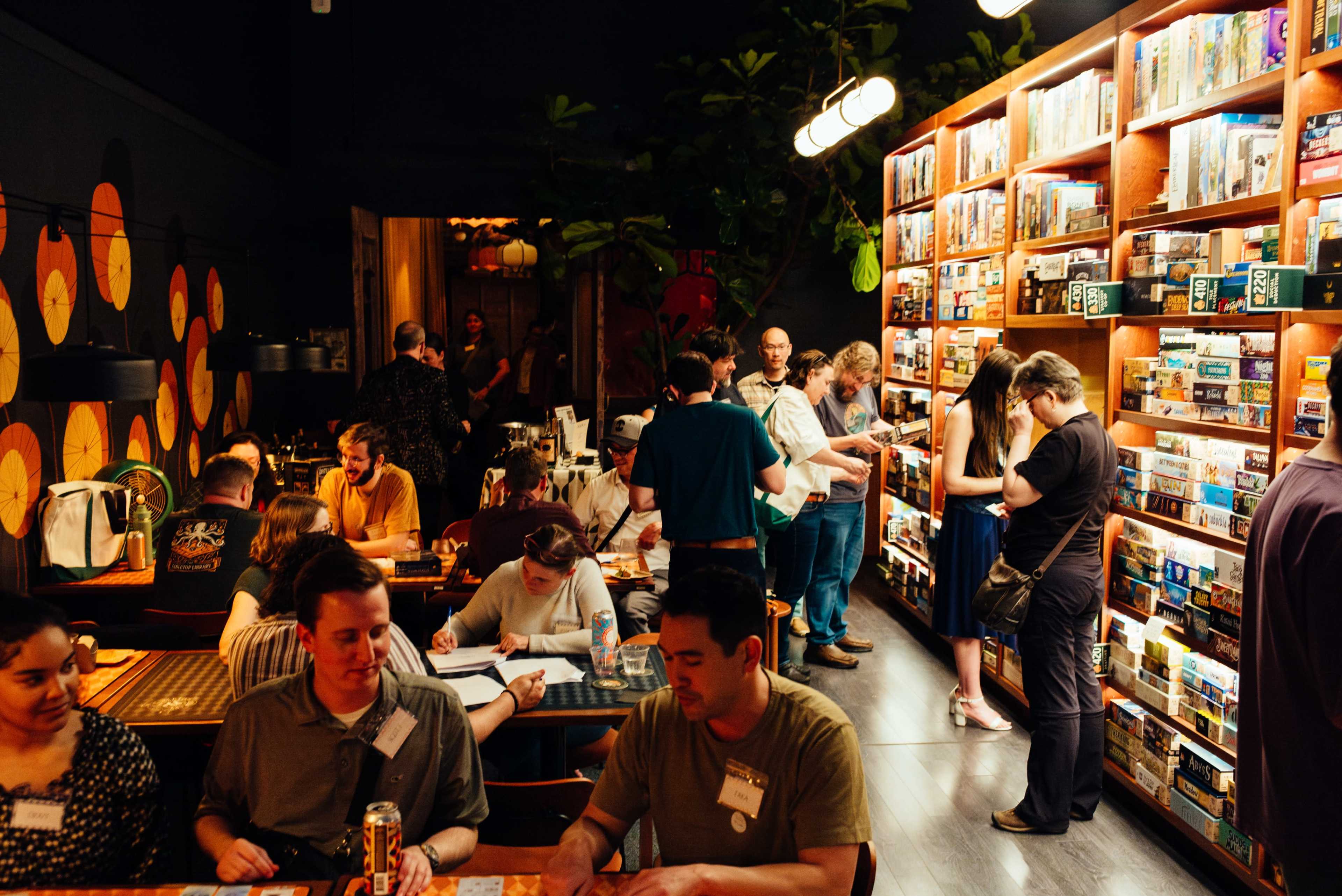 A busy board game café features patrons engaged in games and conversations, with shelves of game boxes lining the walls.