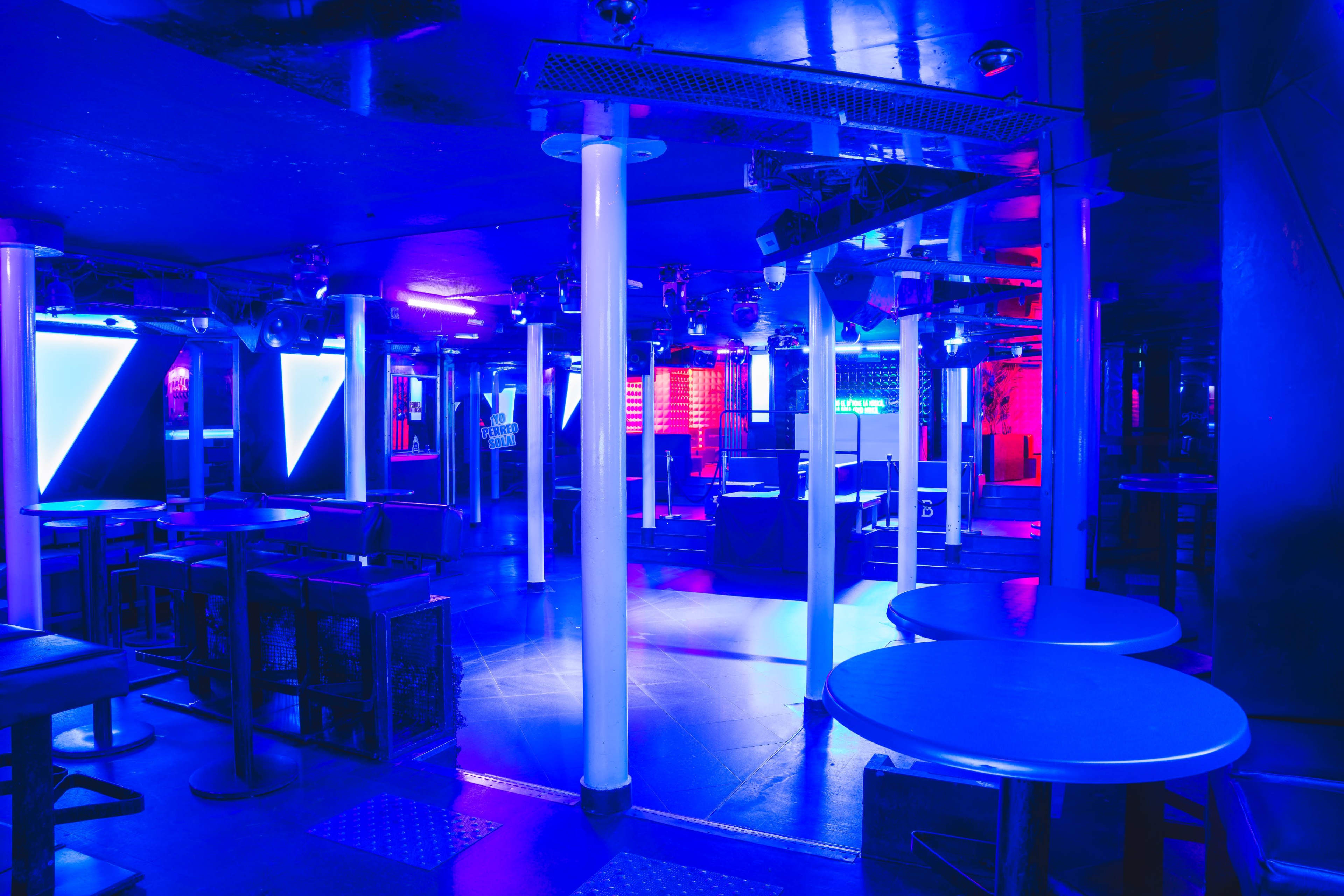 A nightclub interior features blue and purple lighting, empty tables, and a DJ booth at the back.