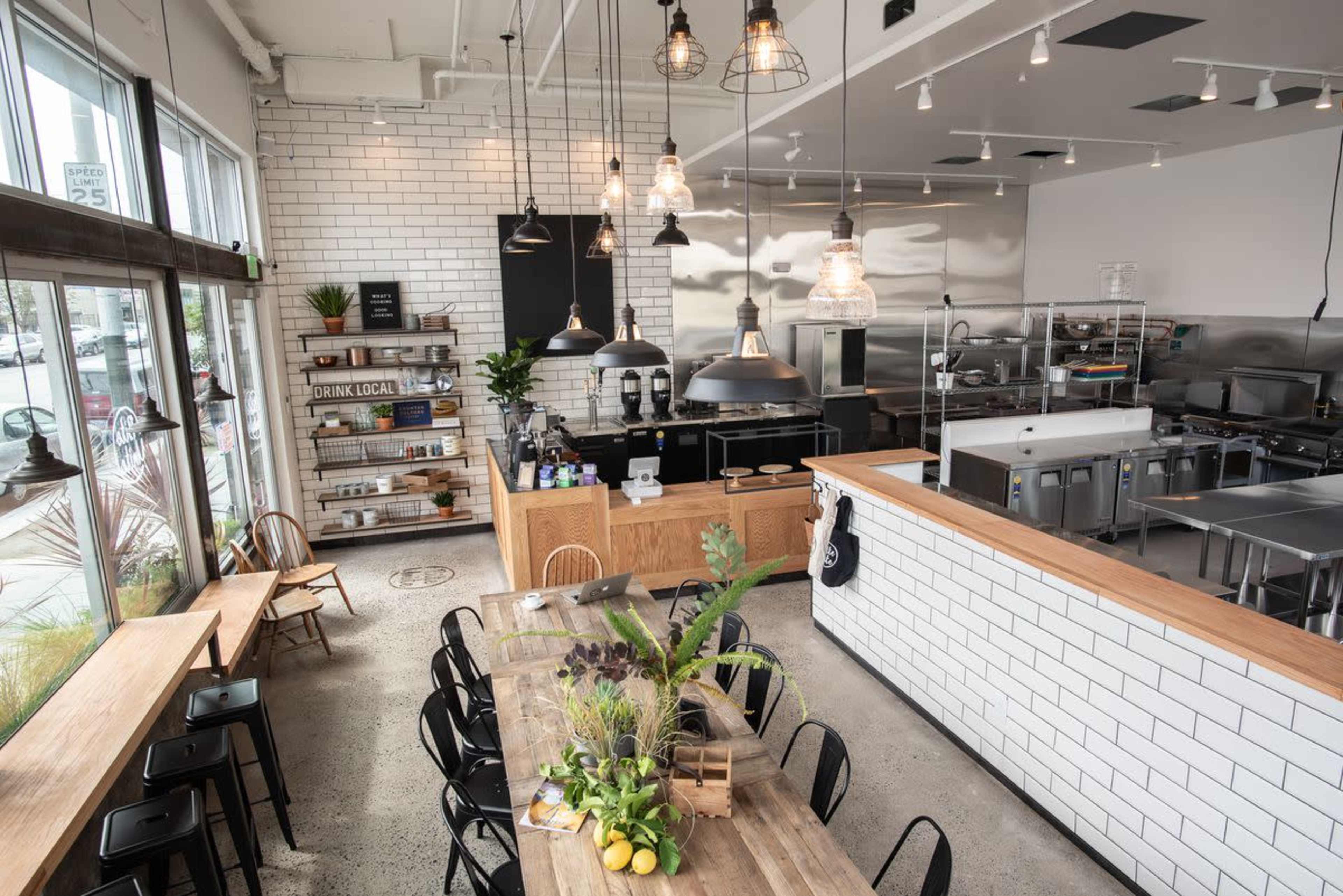 A modern cafe interior with a long wooden table, black chairs, industrial lighting, and a stainless steel kitchen in the background.