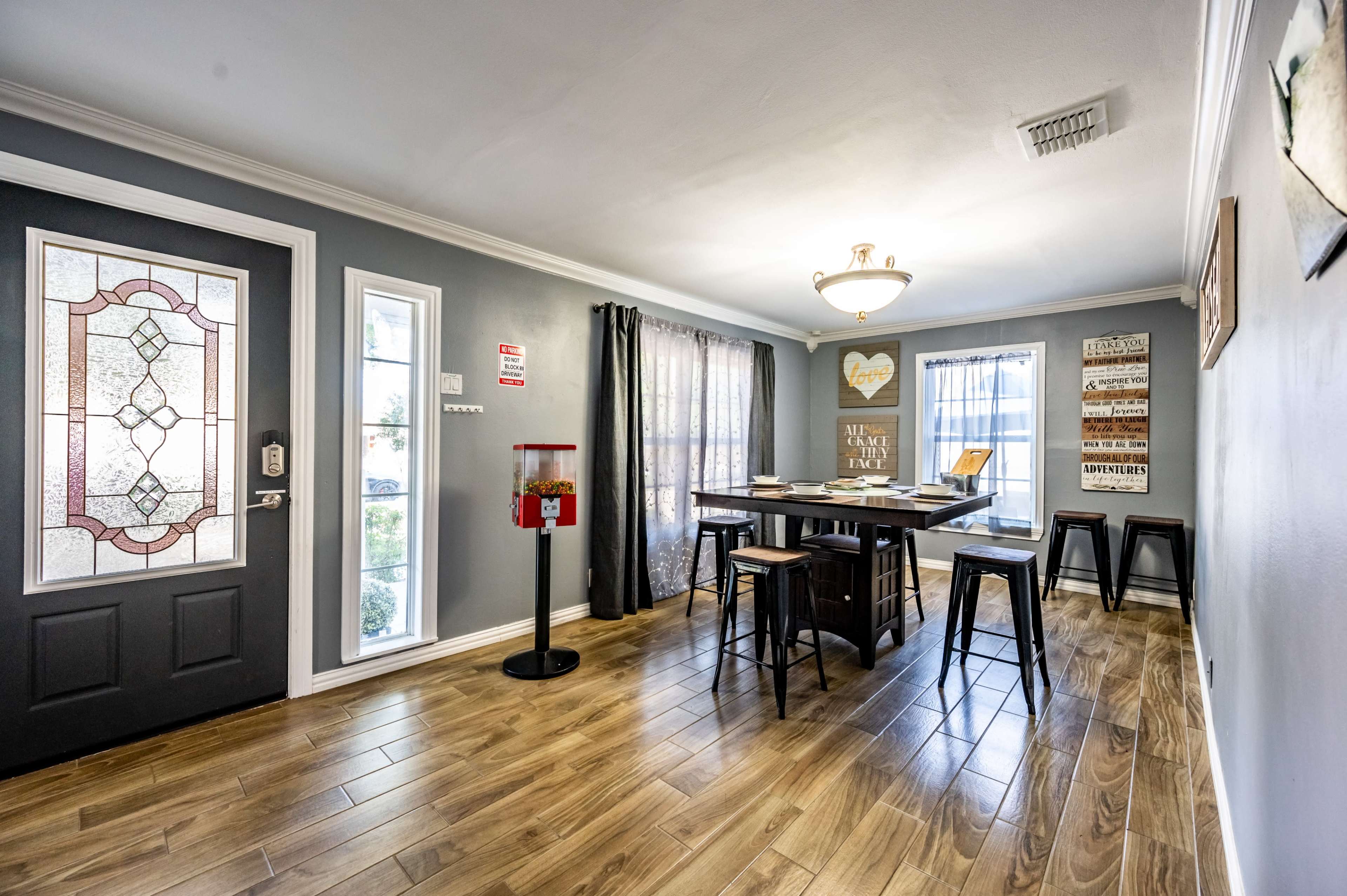 The image shows a dining area featuring a rectangular table surrounded by high stools, with a front door on the left and windows draped with curtains.