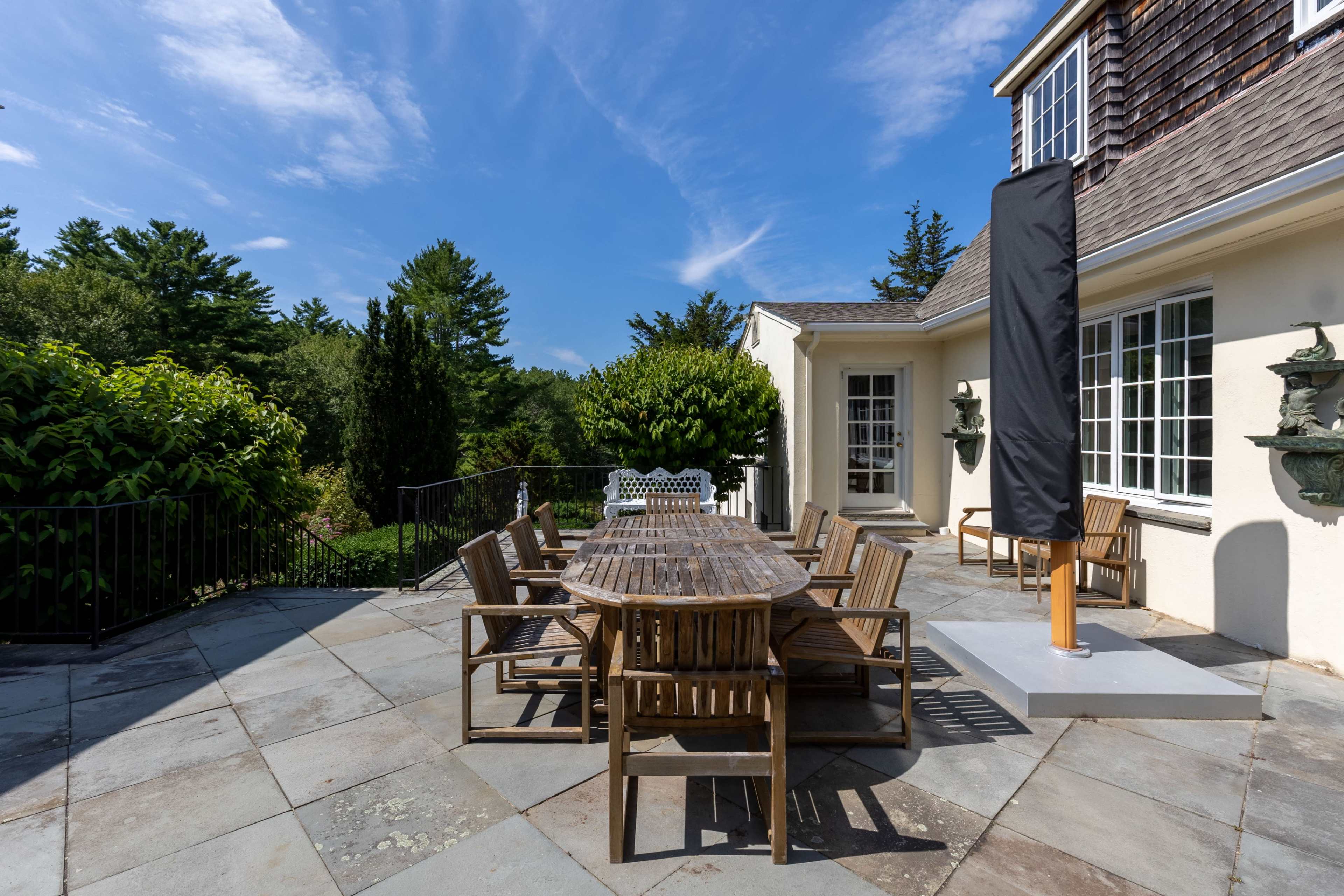 A wooden dining table surrounded by chairs is set on a stone patio with greenery and a house in the background under a blue sky.