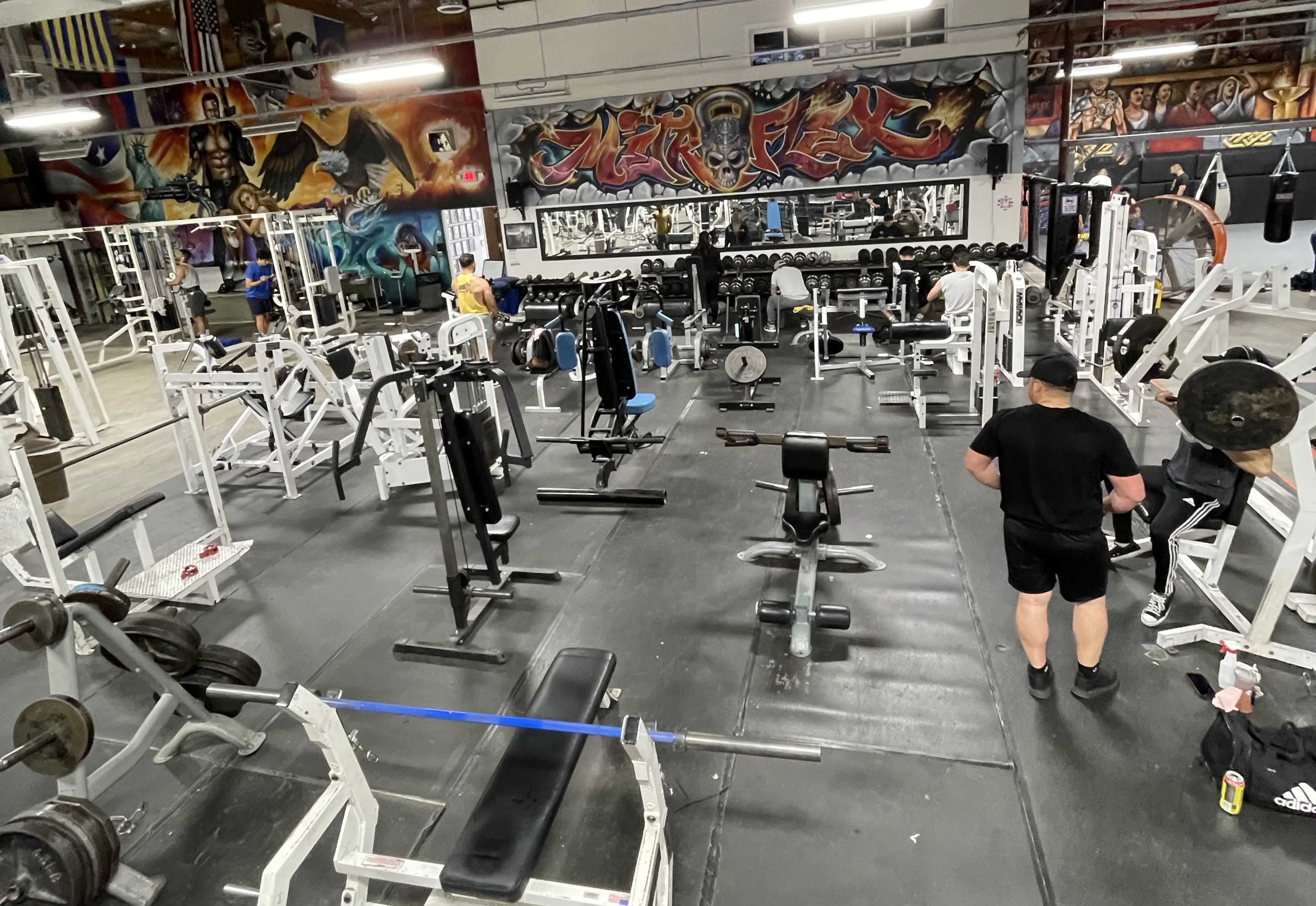A gym filled with various weightlifting equipment and several people engaged in workouts, with colorful murals decorating the walls.