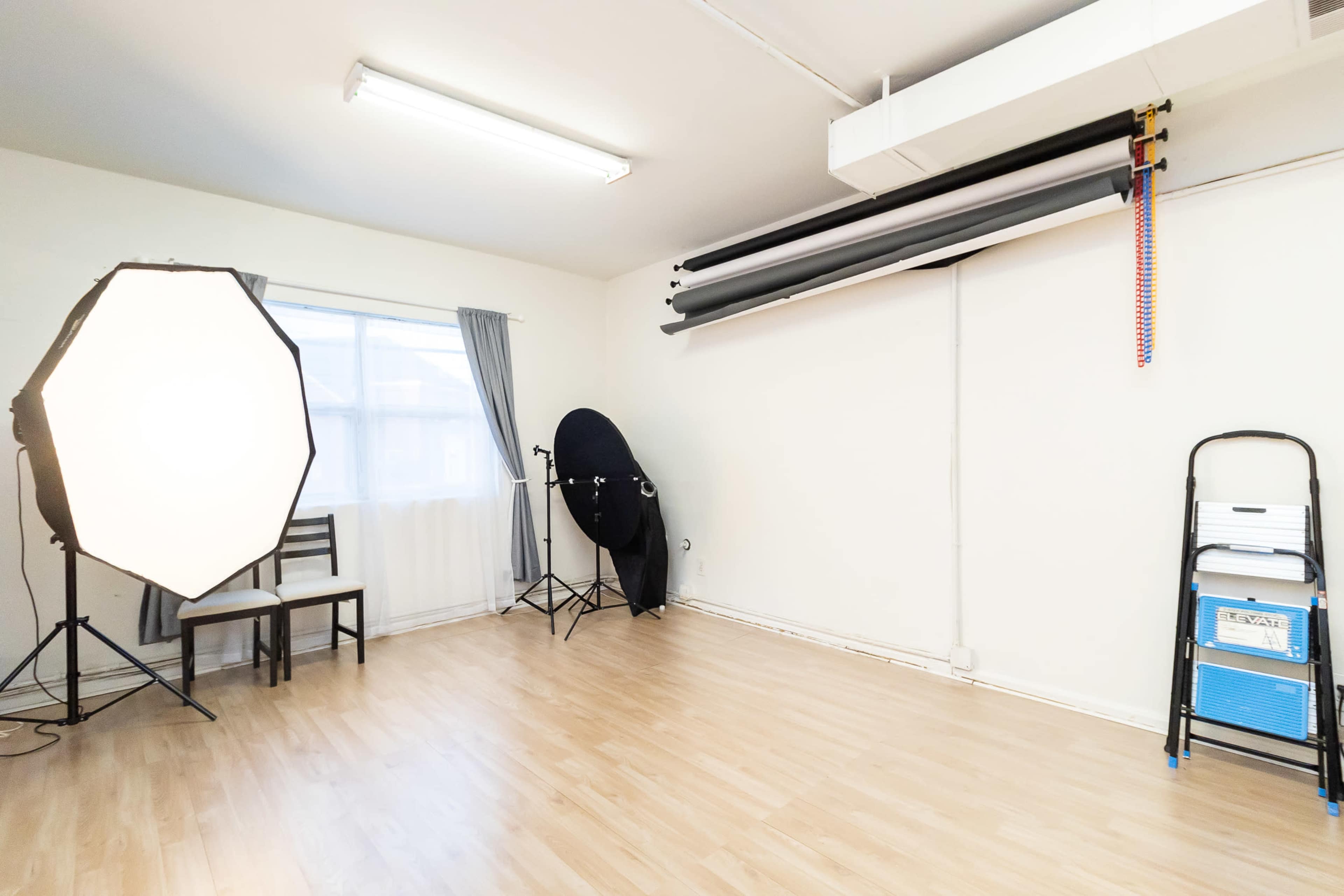 The image shows a minimalistic photography studio with a large softbox, a backdrop setup, a chair, and a step ladder against white walls.
