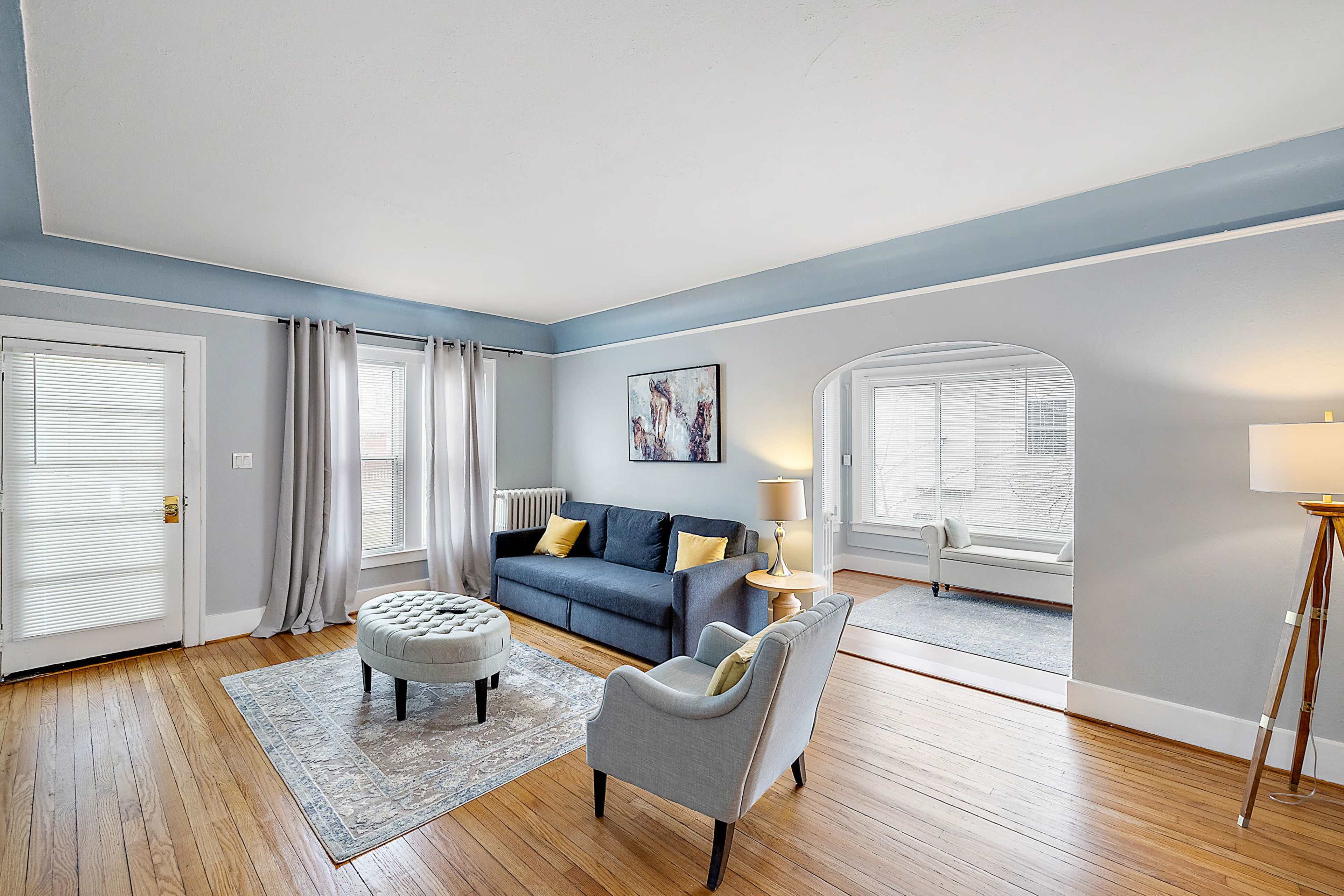 The living room features a blue sofa, a round ottoman, and a chair, with large windows allowing natural light to fill the space.