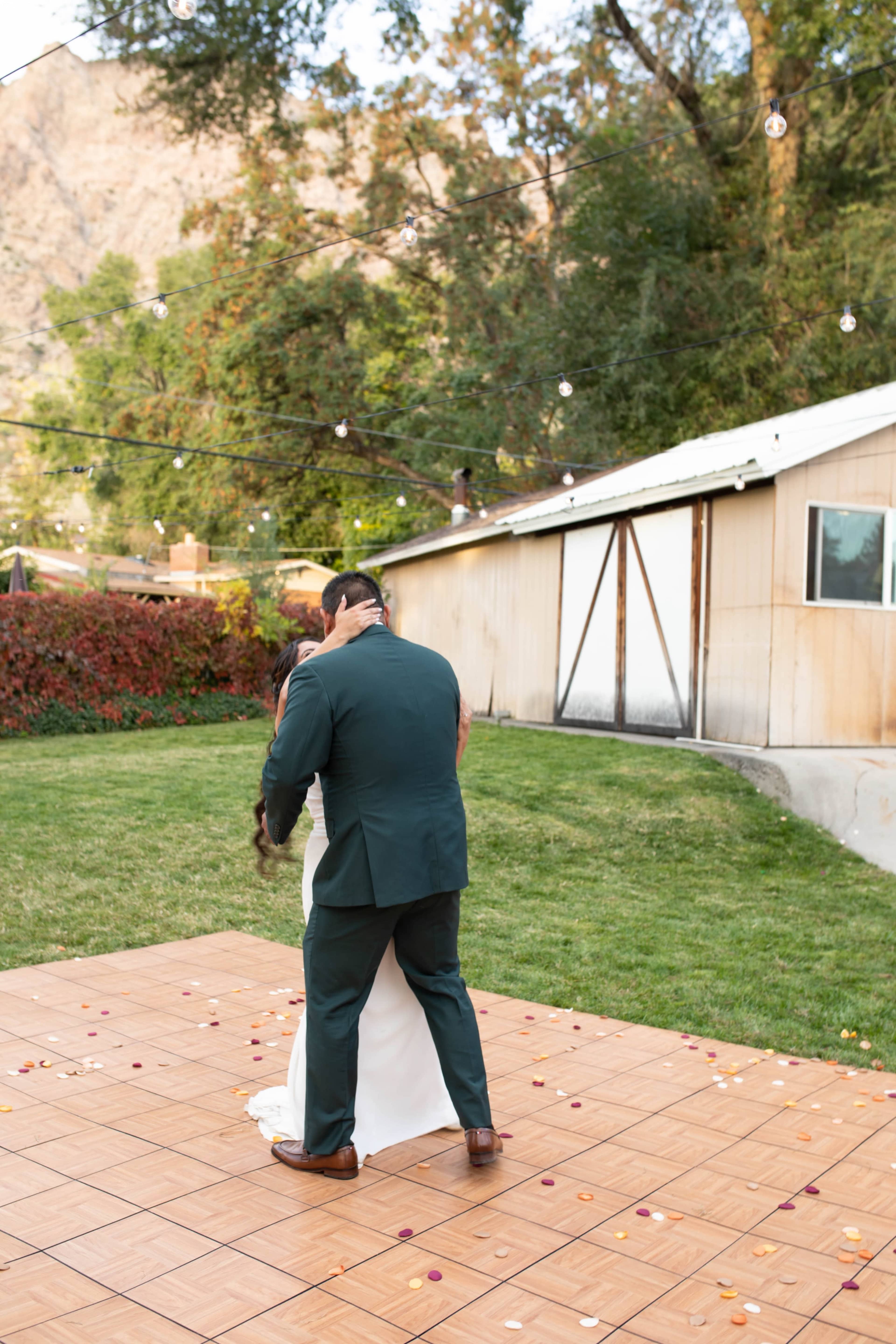 A couple shares a kiss on a dance floor surrounded by greenery and a barn in the background, with string lights overhead.