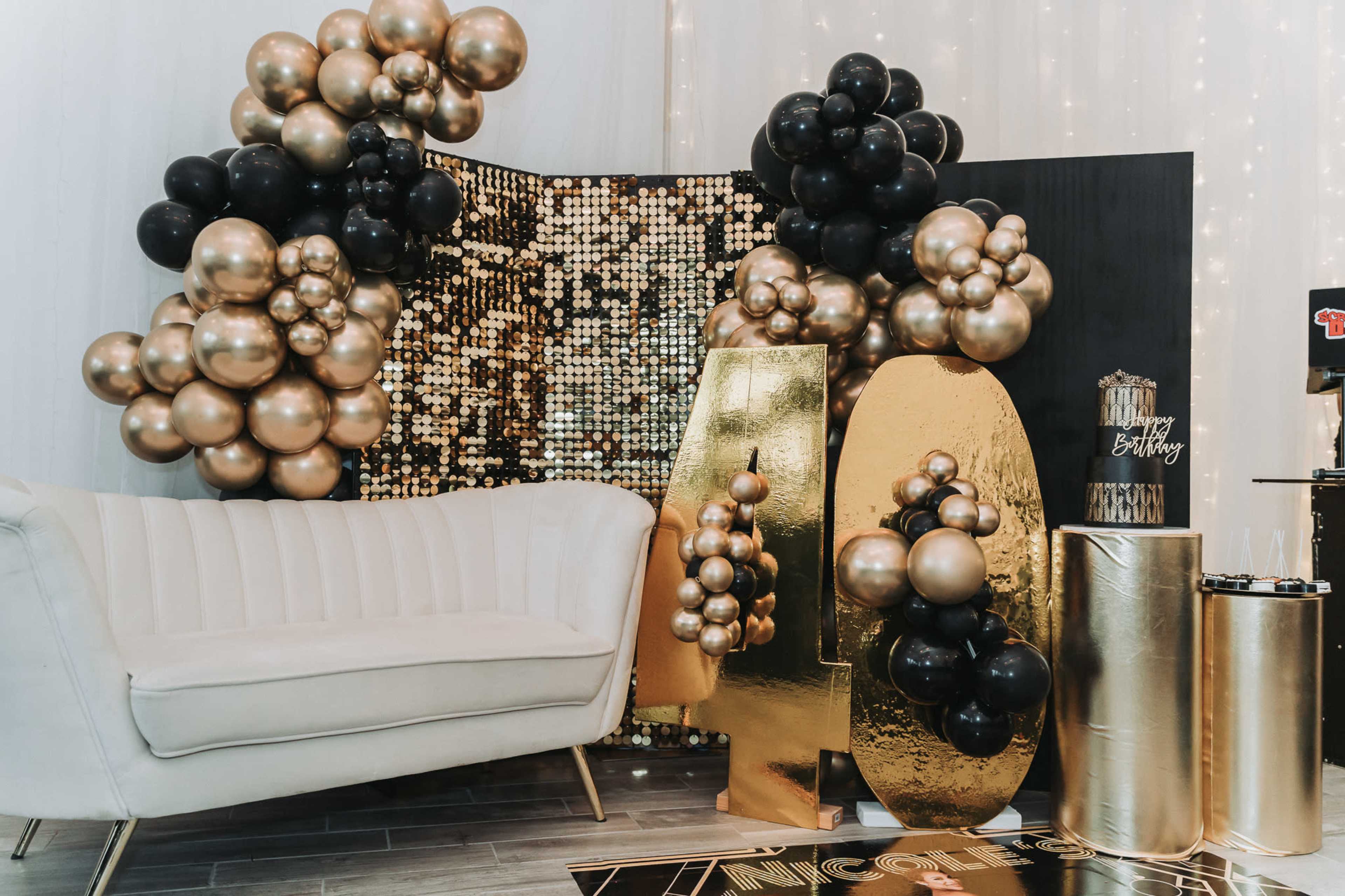 A festive birthday party setup with a white couch, large gold and black balloons, and a backdrop of shimmering sequins.