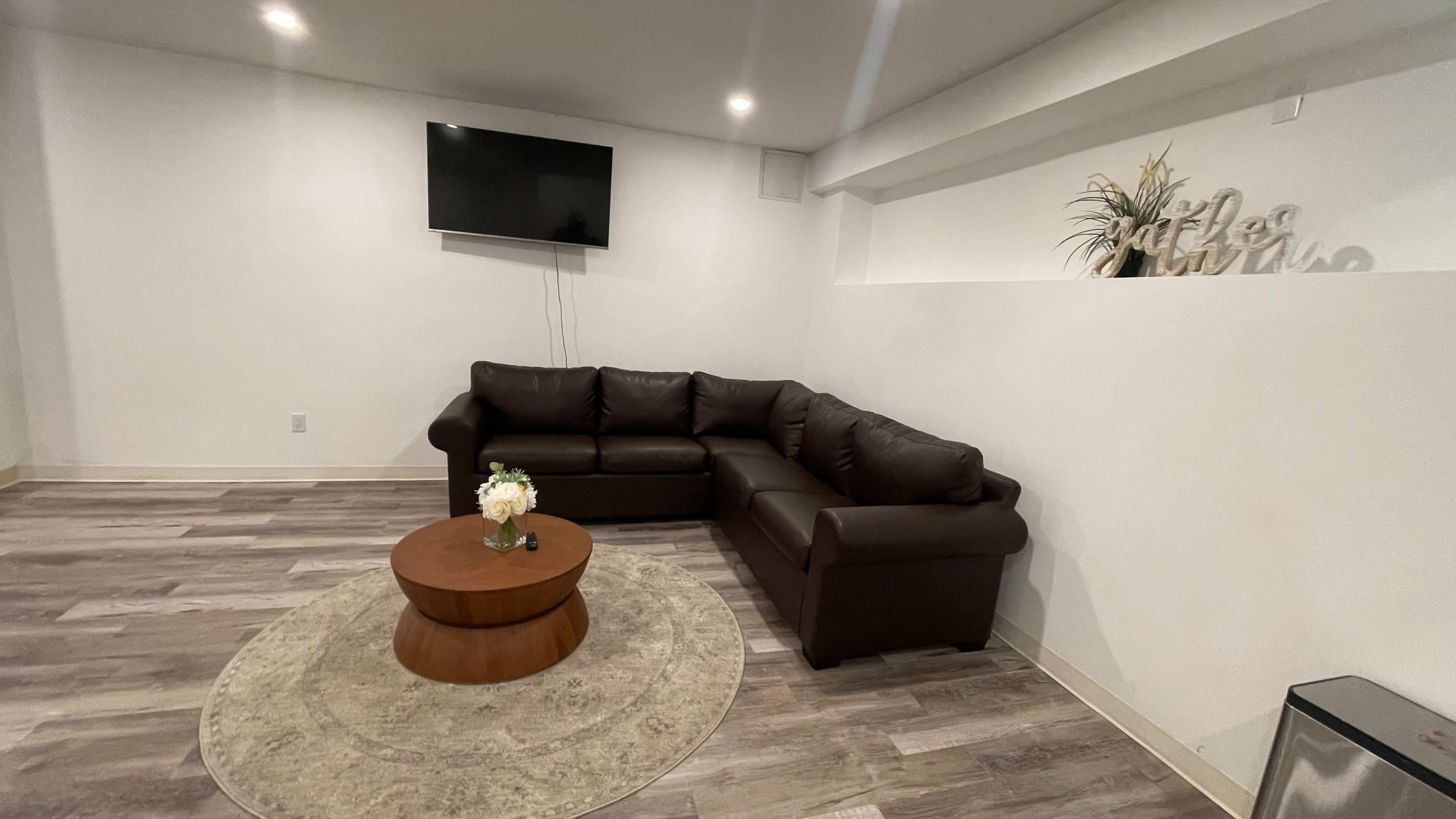 A modern living room with a brown sectional sofa, a round wooden coffee table, and a television mounted on the wall.