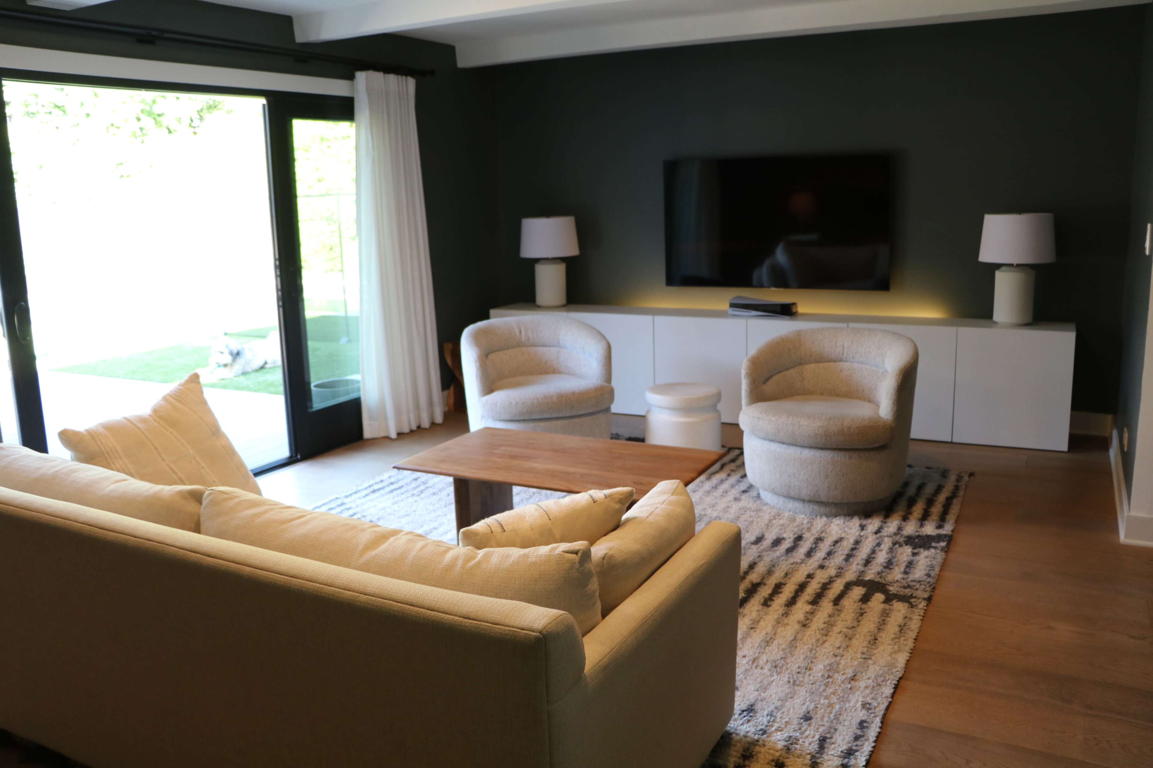 A modern living room features a beige sofa, two curved chairs, a wooden coffee table, and a TV mounted on a dark wall, with large glass doors opening to a garden.