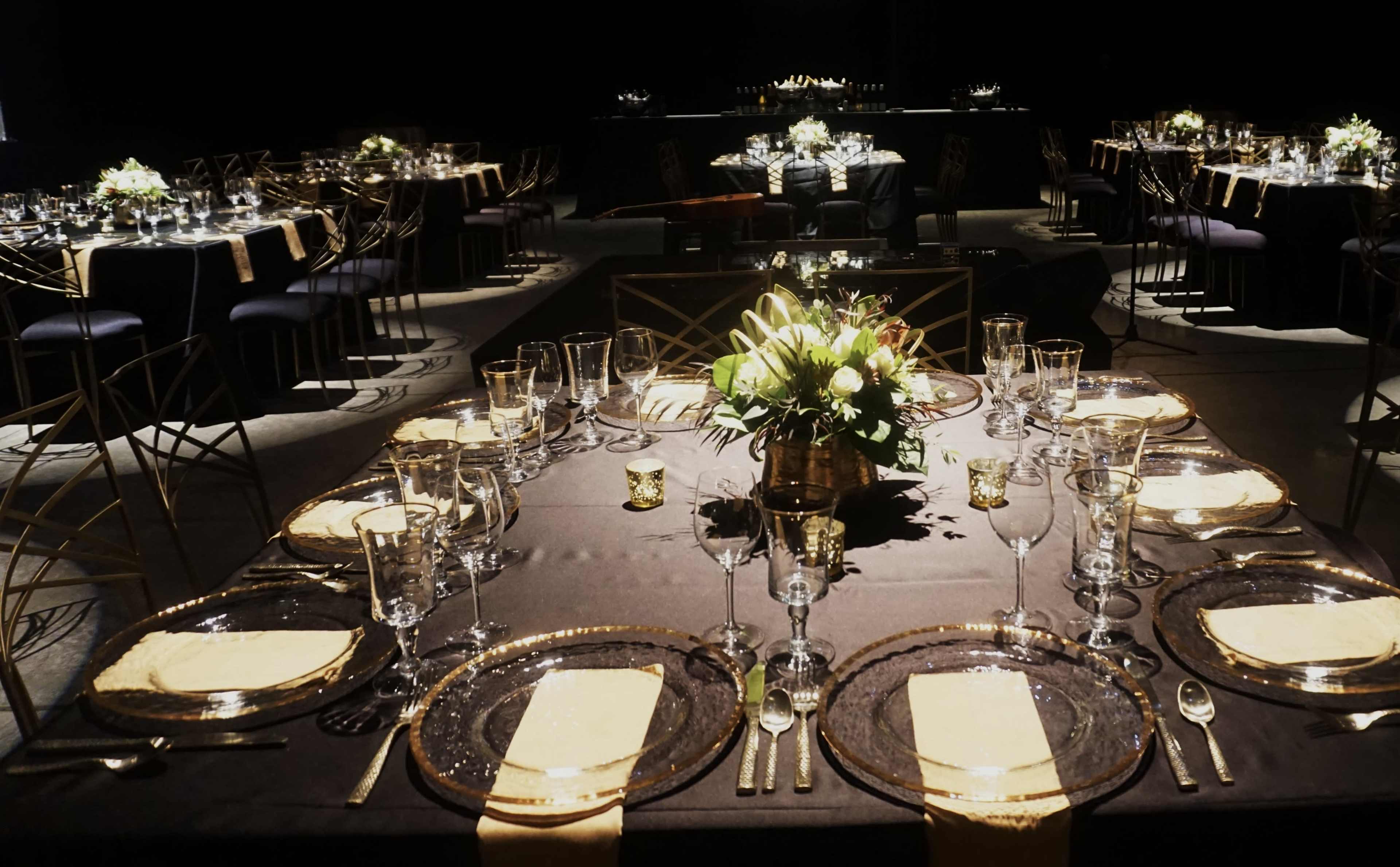 The image shows a dimly lit banquet setting with elegantly arranged tables featuring dark tablecloths, glassware, and floral centerpieces.
