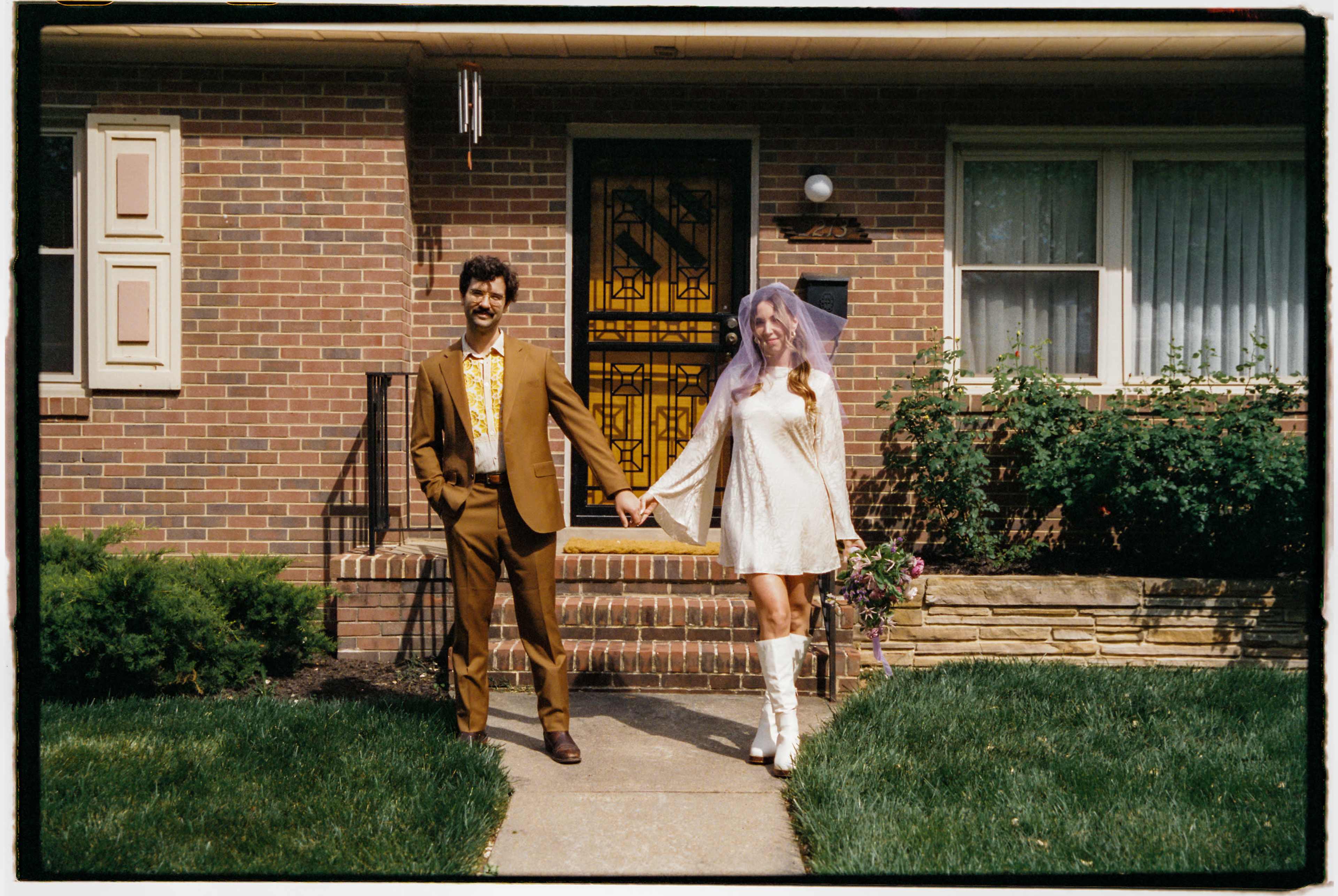 A couple stands in front of a brick house, holding hands, with the man in a brown suit and the woman in a white dress and boots, wearing a veil.