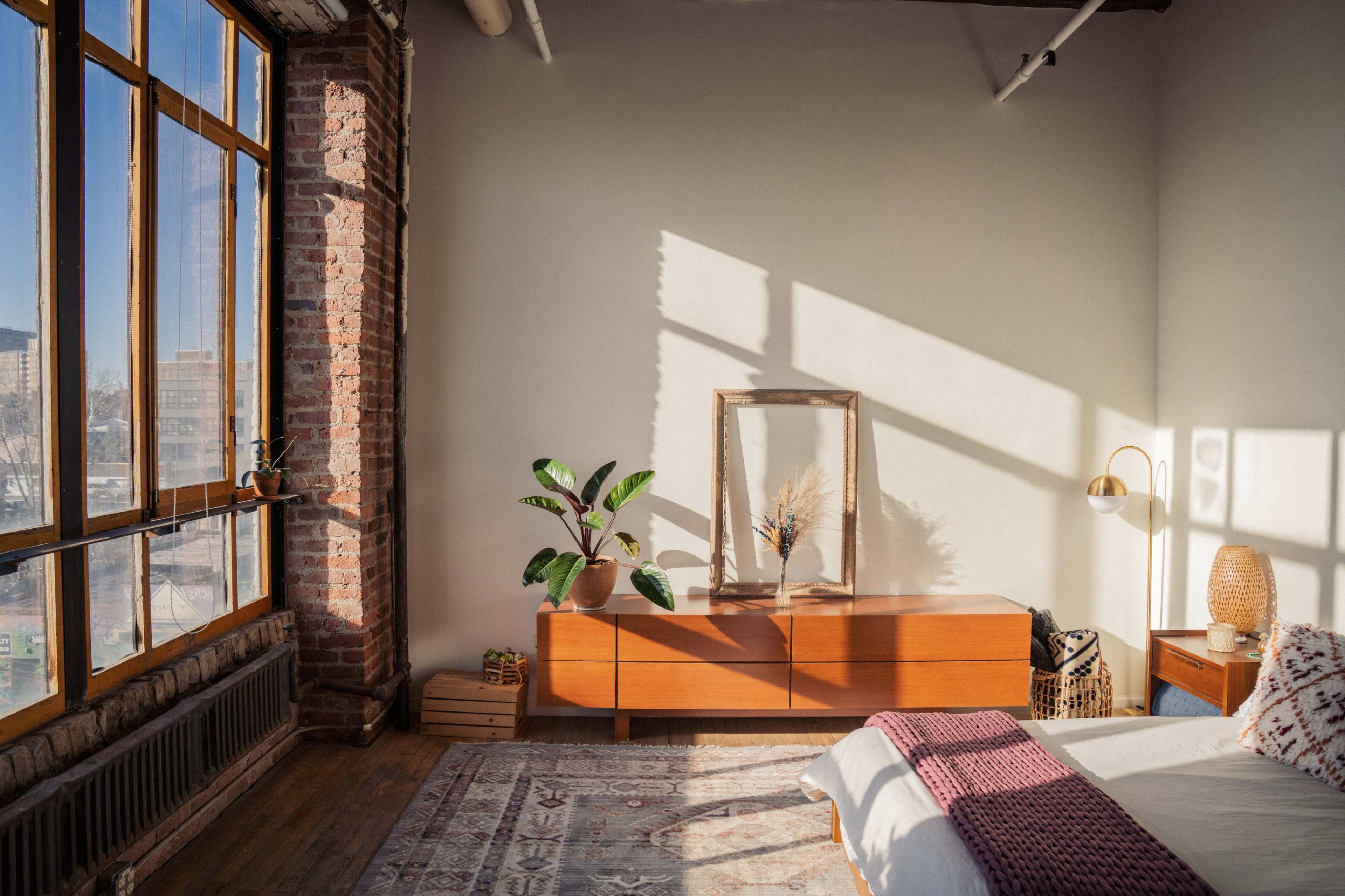 A sunlit bedroom features a bed, a wooden dresser, a framed mirror, and a plant, with large windows showcasing a brick wall.