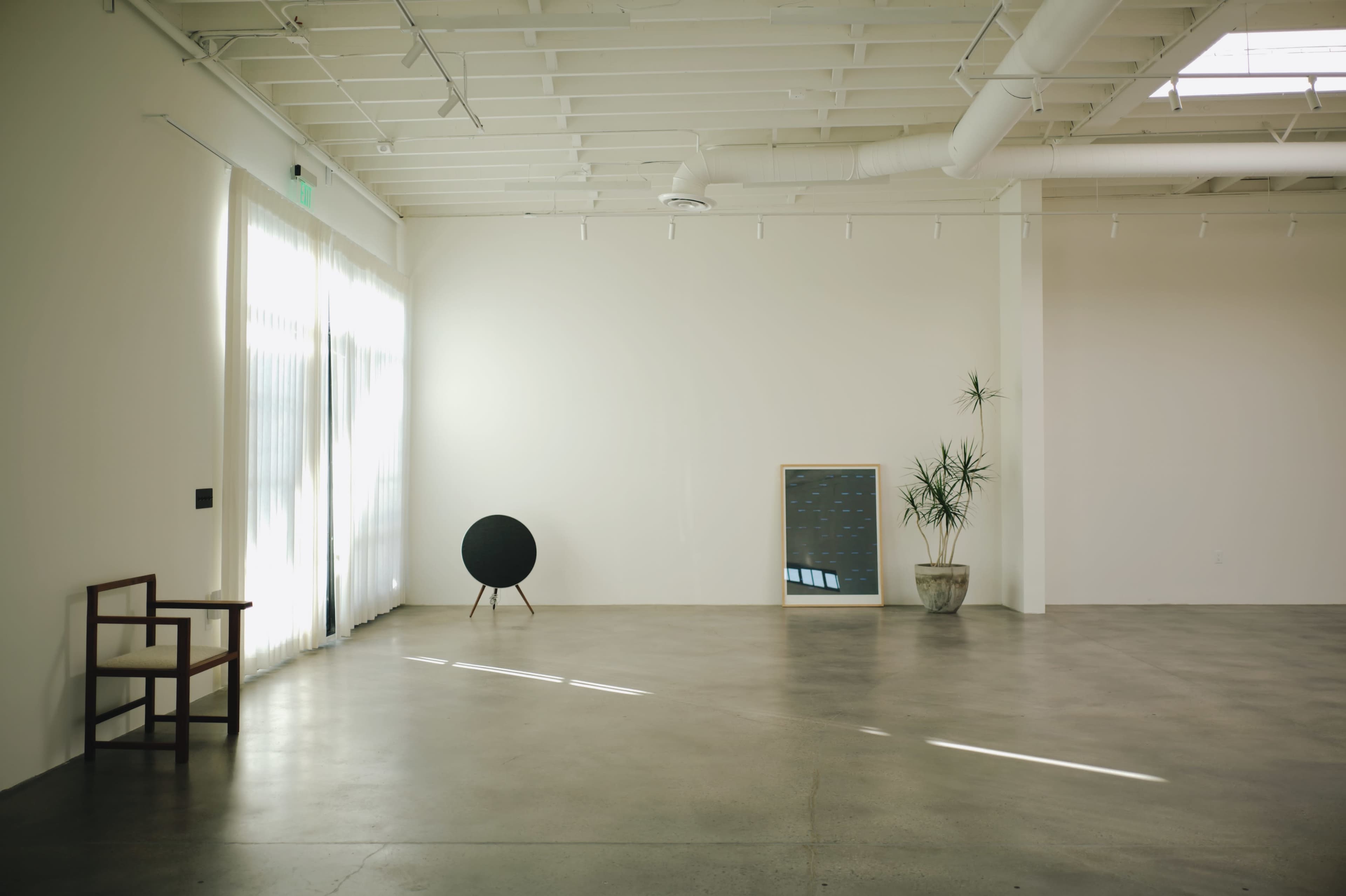 A spacious, minimalist room features a wooden chair, a round black object on a stand, a potted plant, and a mirror against a white wall.