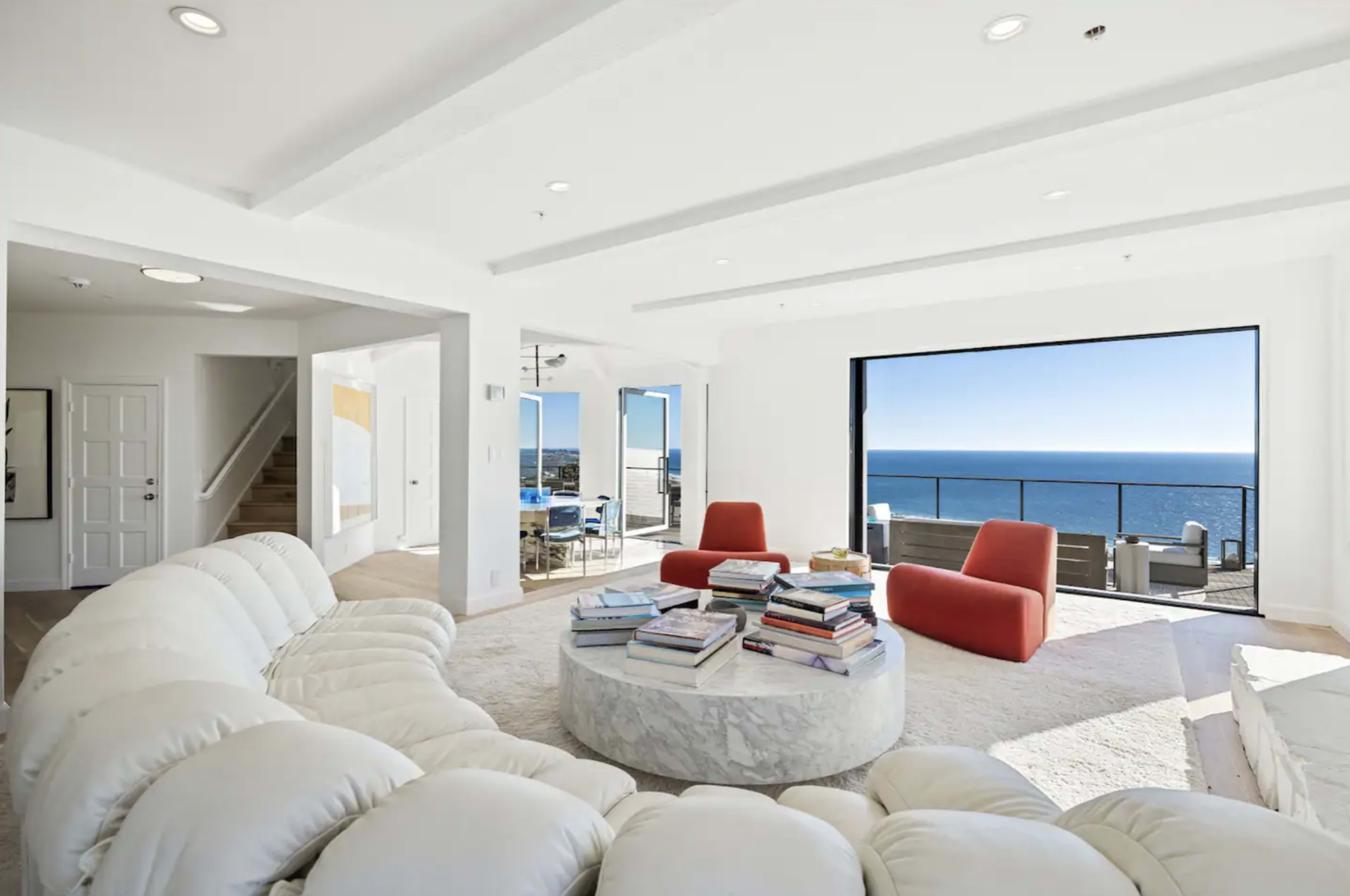 A modern living room features a circular sofa, a marble coffee table stacked with books, large windows overlooking the ocean, and two accent chairs.