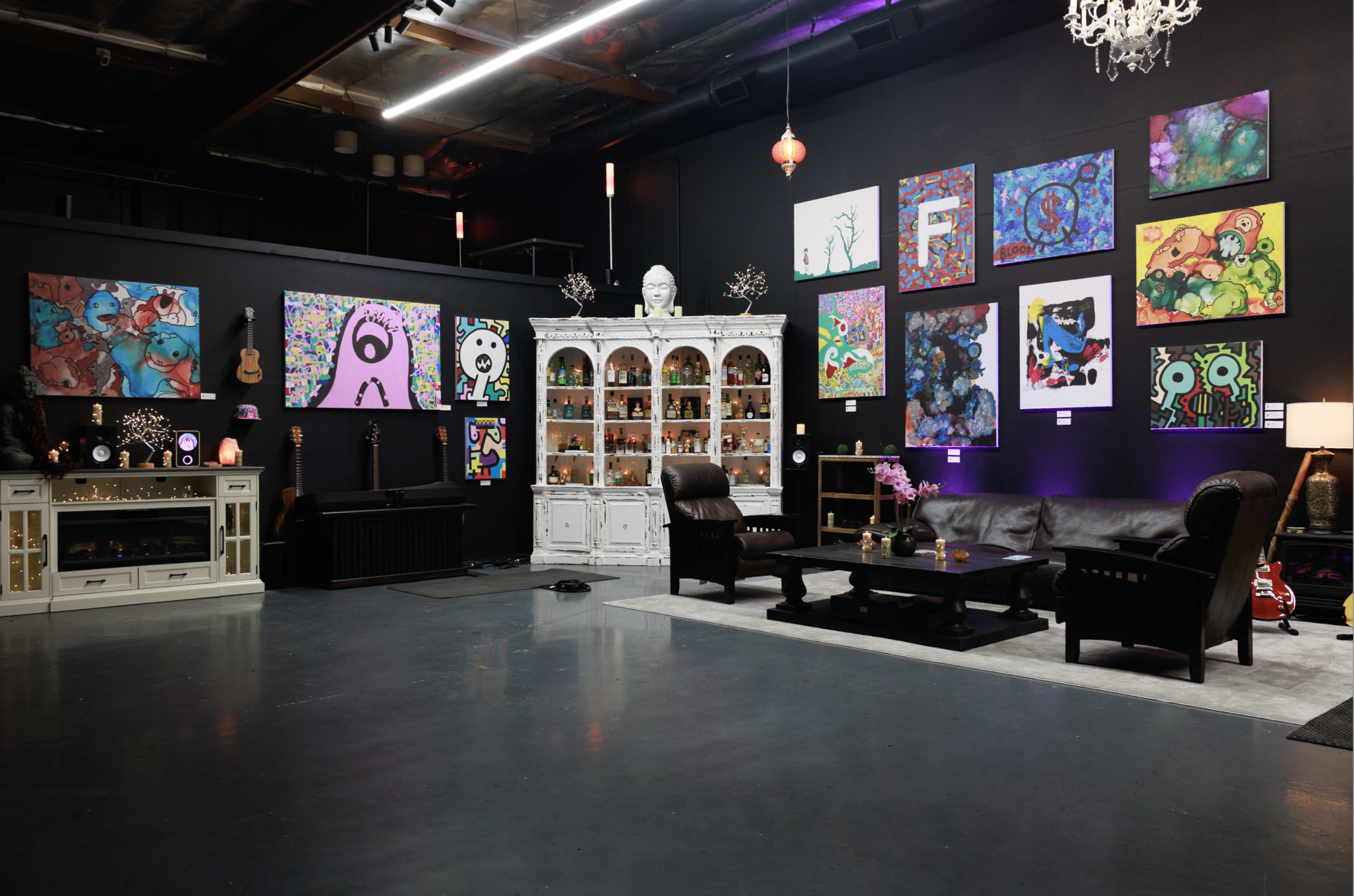 The scene features a modern art studio with vibrant paintings on the black walls, a white display cabinet, two dark chairs, a coffee table, and decorative lighting.