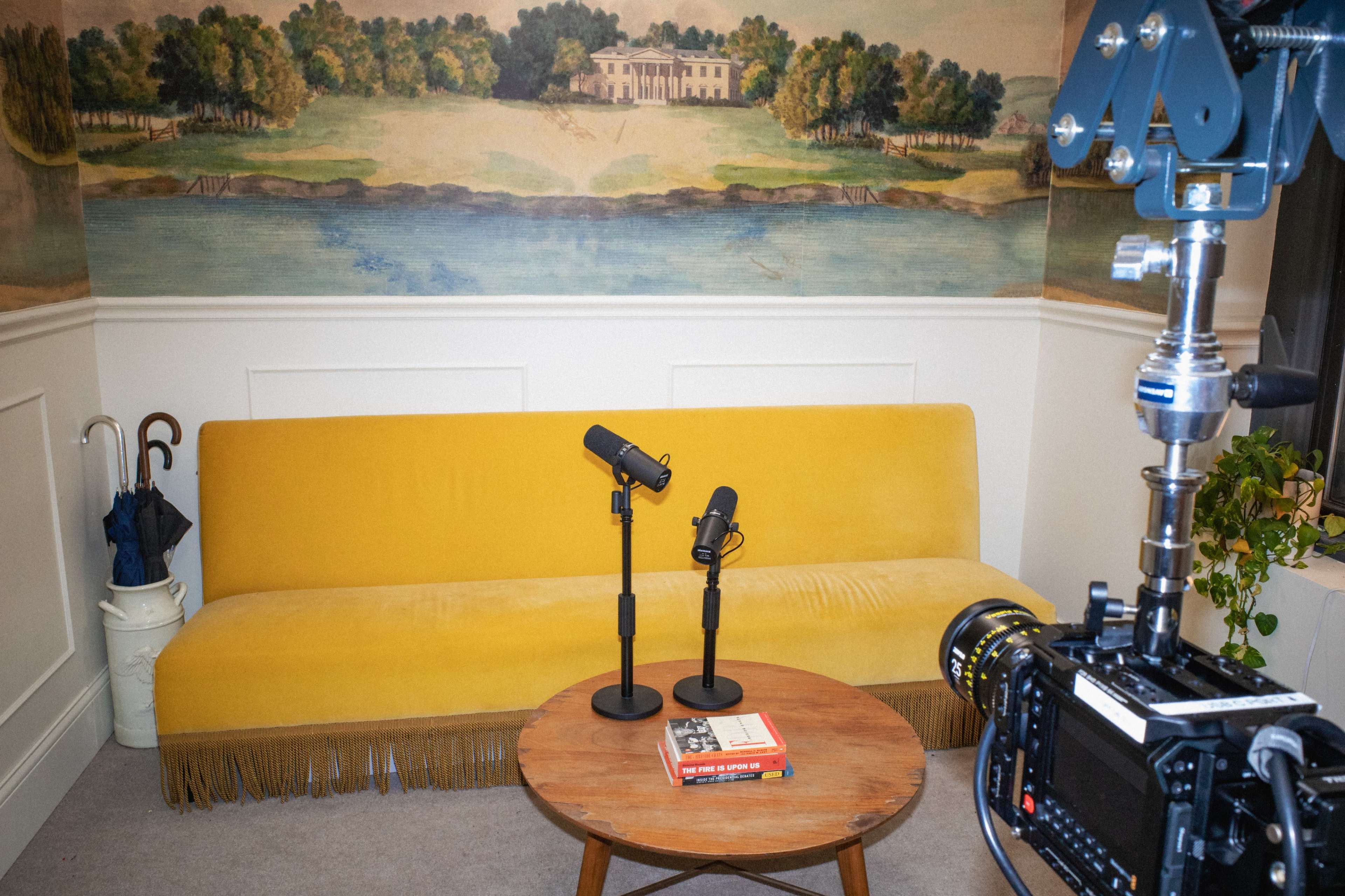 A yellow sofa with fringe is positioned in front of a painted landscape mural, accompanied by two microphones on stands and a wooden coffee table with a book, while a camera rig is set up nearby.