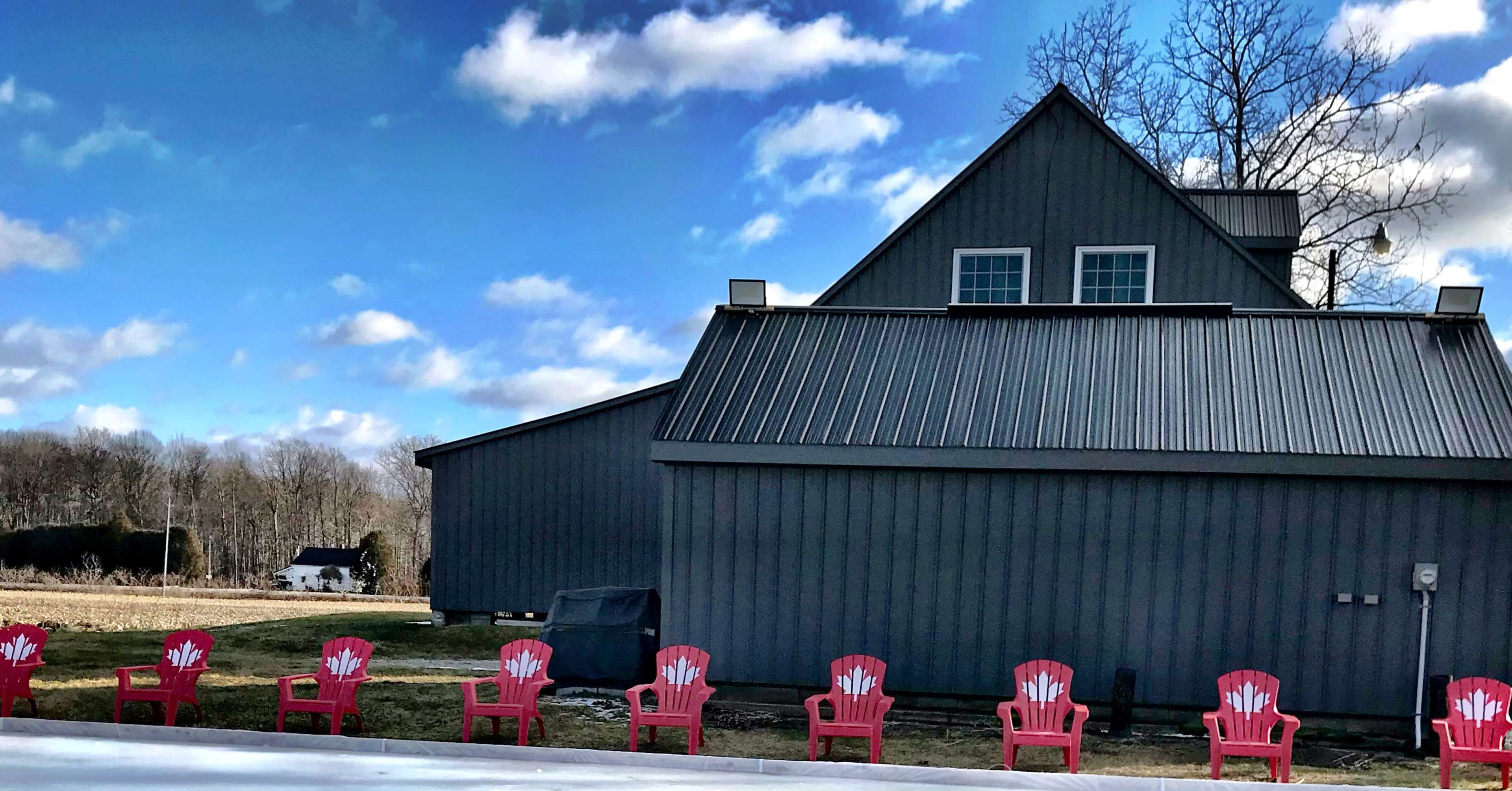 A row of red Adirondack chairs is positioned in front of a gray barn-style building under a partly cloudy sky.