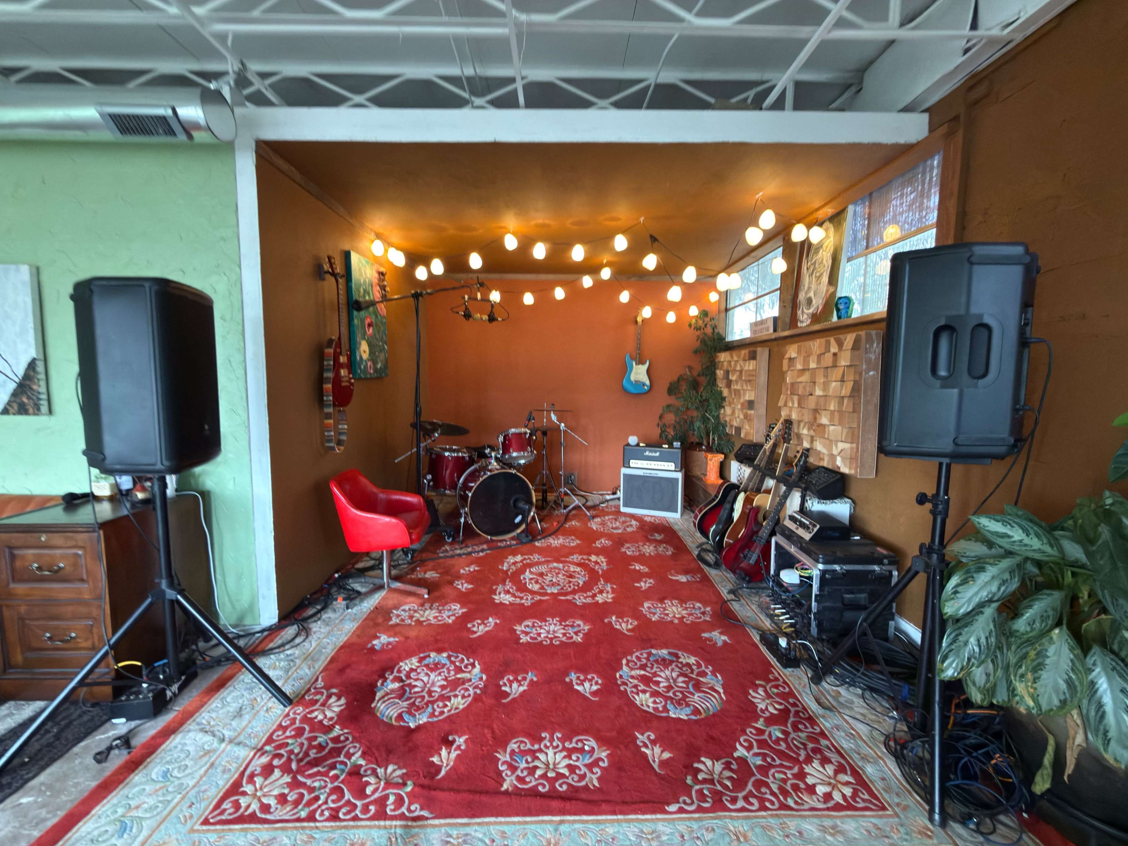 The image shows a small music performance space with a red carpet, musical instruments, and decorative string lights hanging from the ceiling.