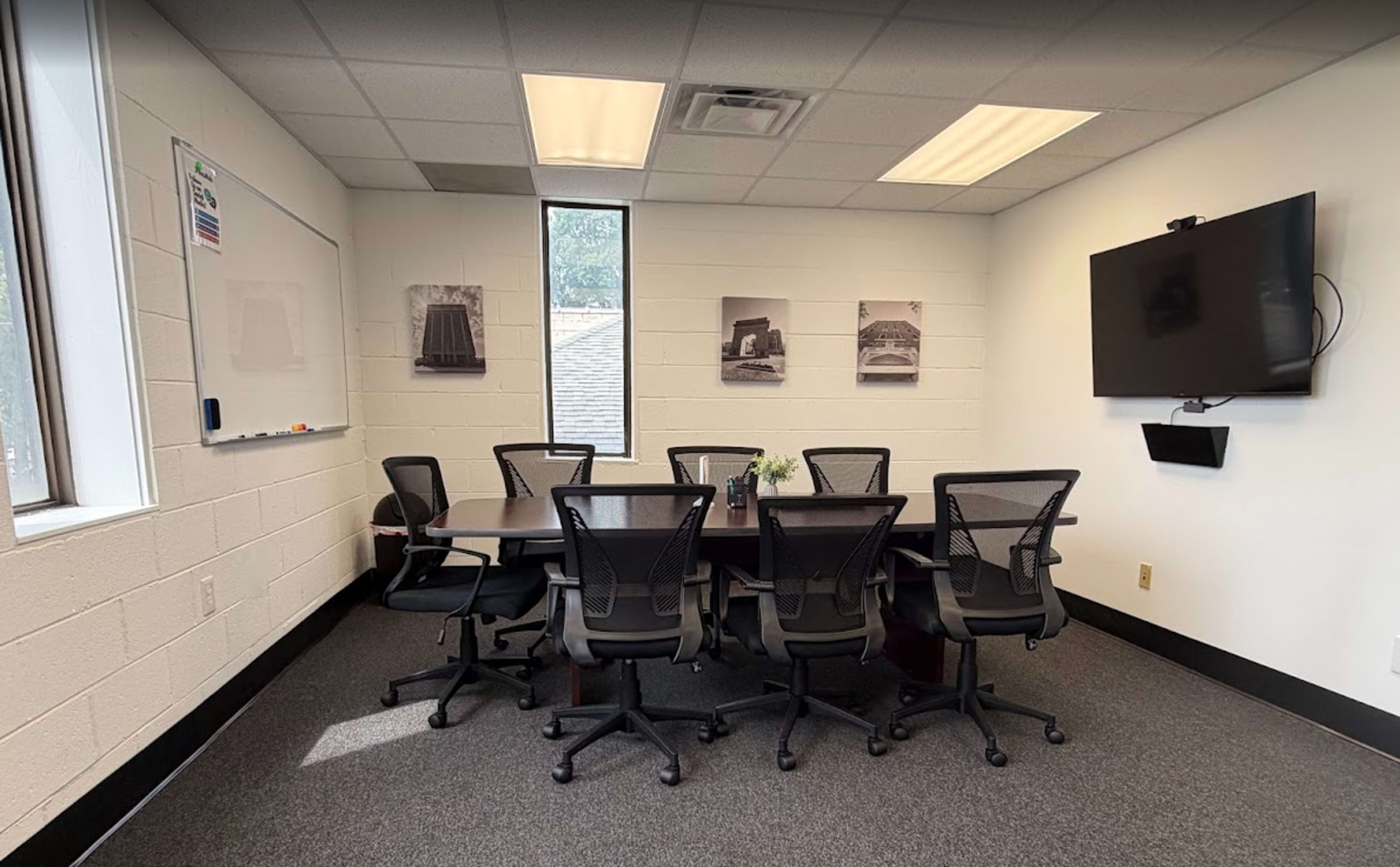 The image shows a conference room with a large table surrounded by eight chairs, a whiteboard on one wall, a television mounted on another, and three framed photographs of buildings on the walls.