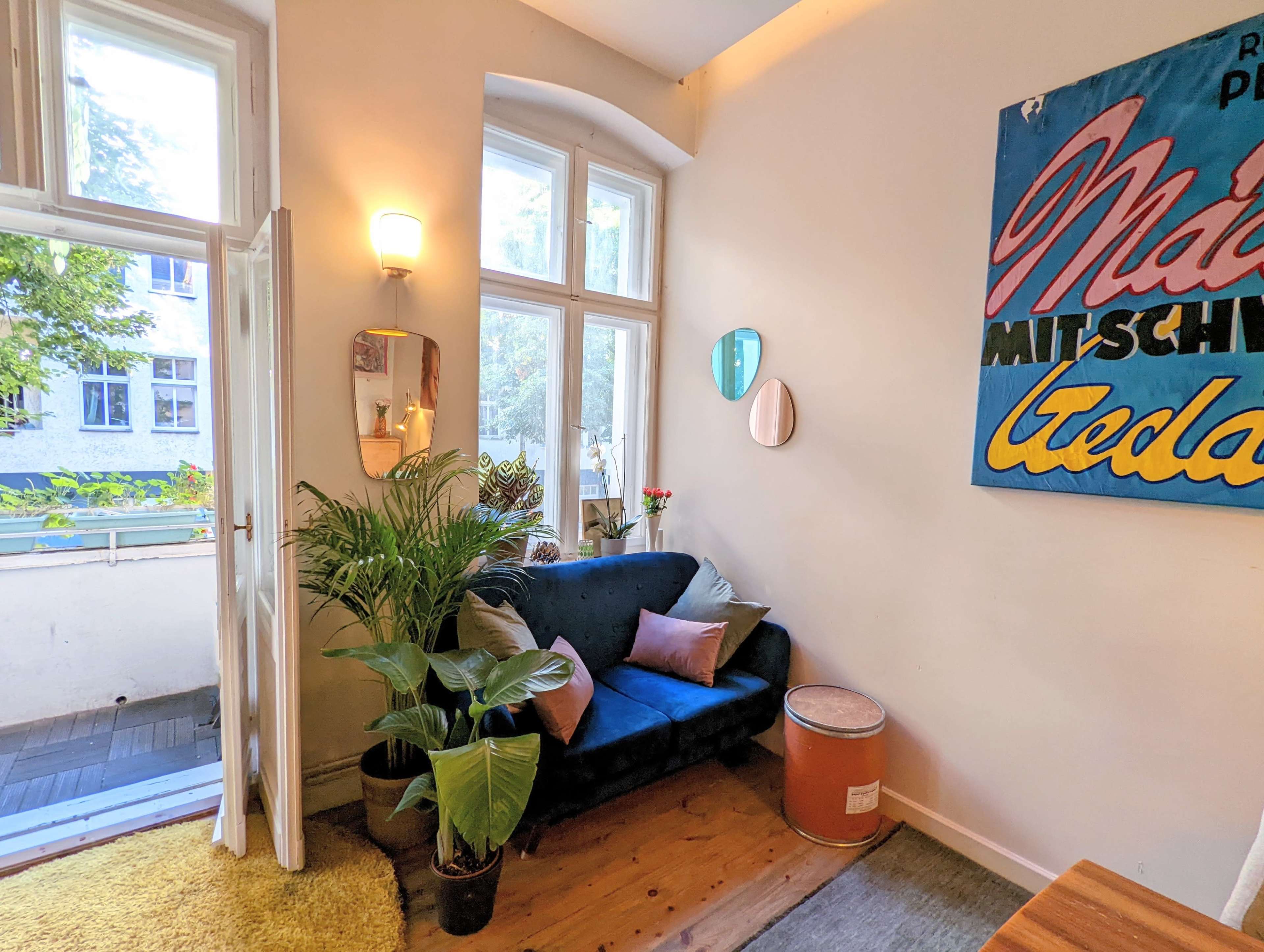 A cozy room featuring a blue sofa, a plant, a small side table, and large windows with a view of greenery outside.