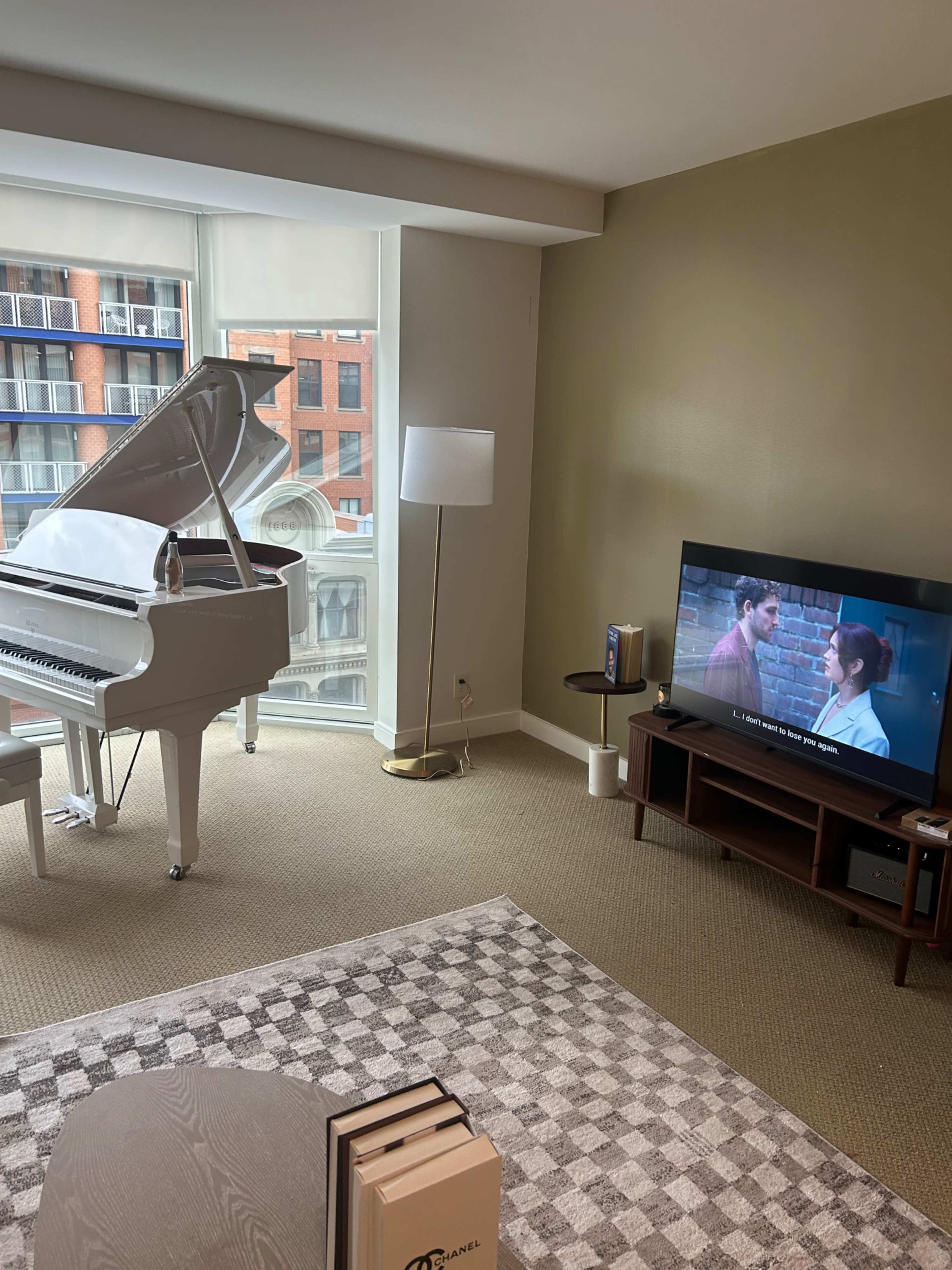 The image shows a modern living room featuring a white piano, a television displaying a scene from a movie, a floor lamp, and a coffee table on a patterned rug.