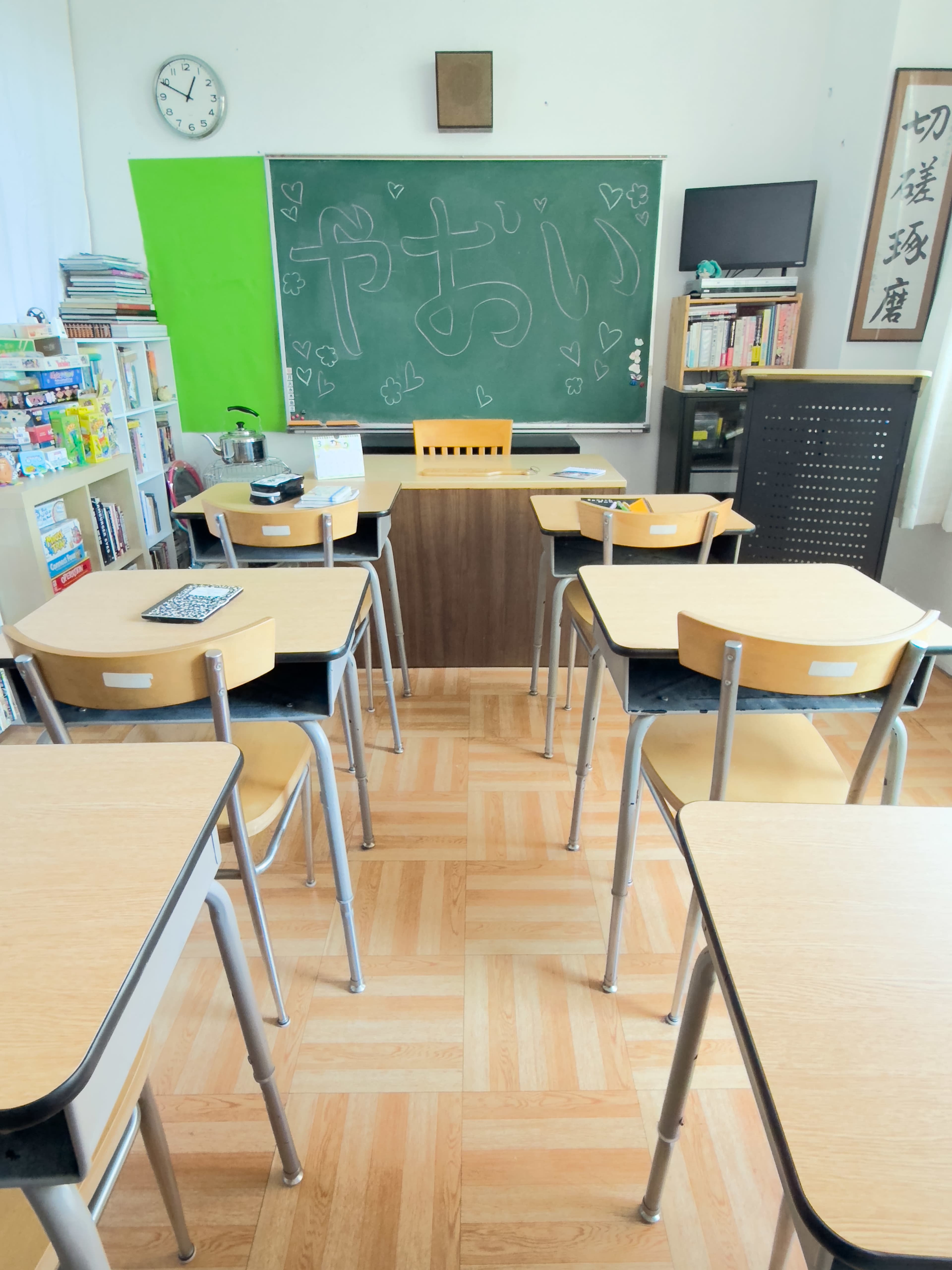 A classroom is arranged with wooden desks and chairs facing a blackboard, which has writing on it, surrounded by shelves filled with books and materials.