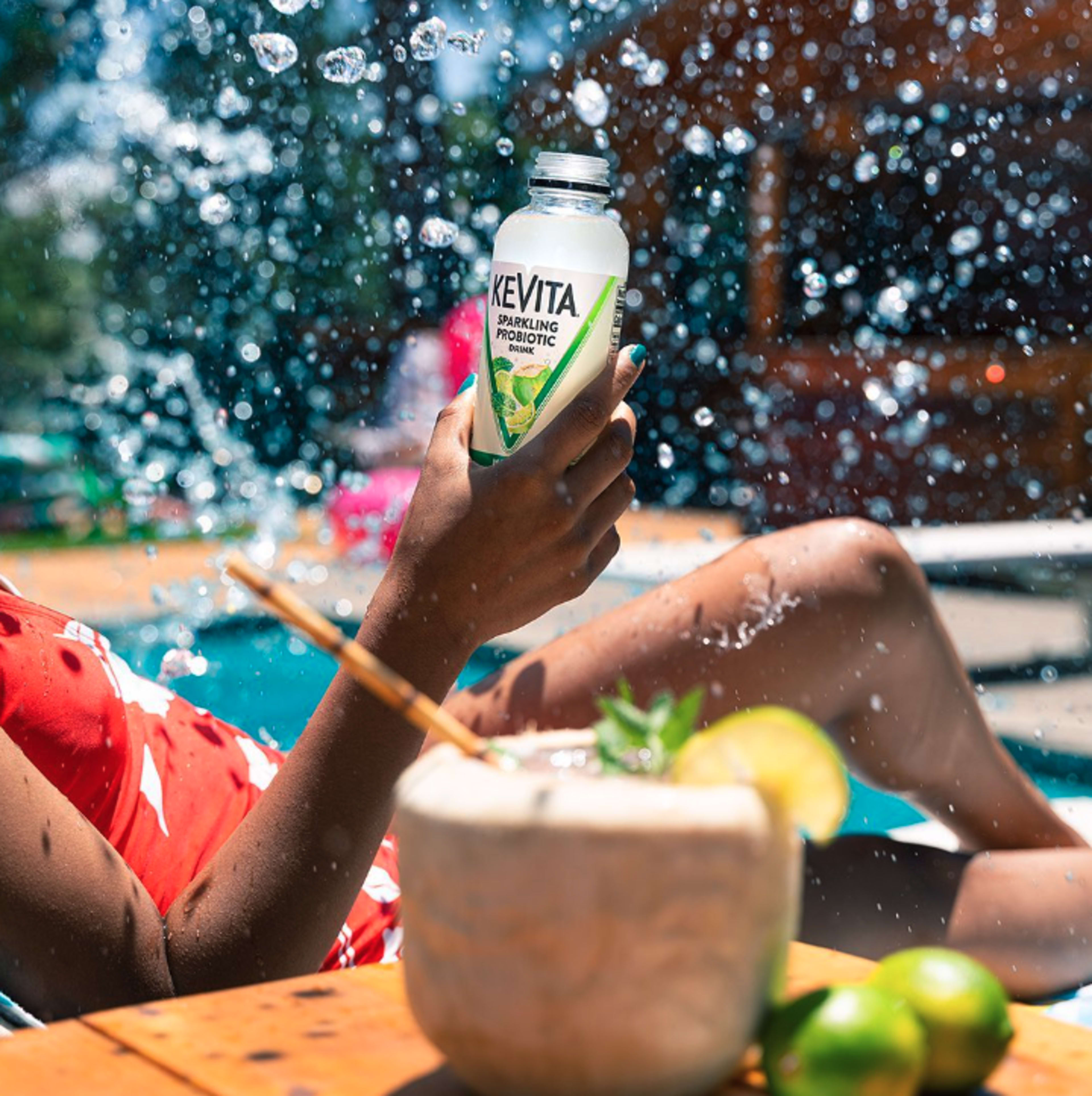 A person in a red swimsuit holds a bottle of Kevita near a pool, with water splashing around them.