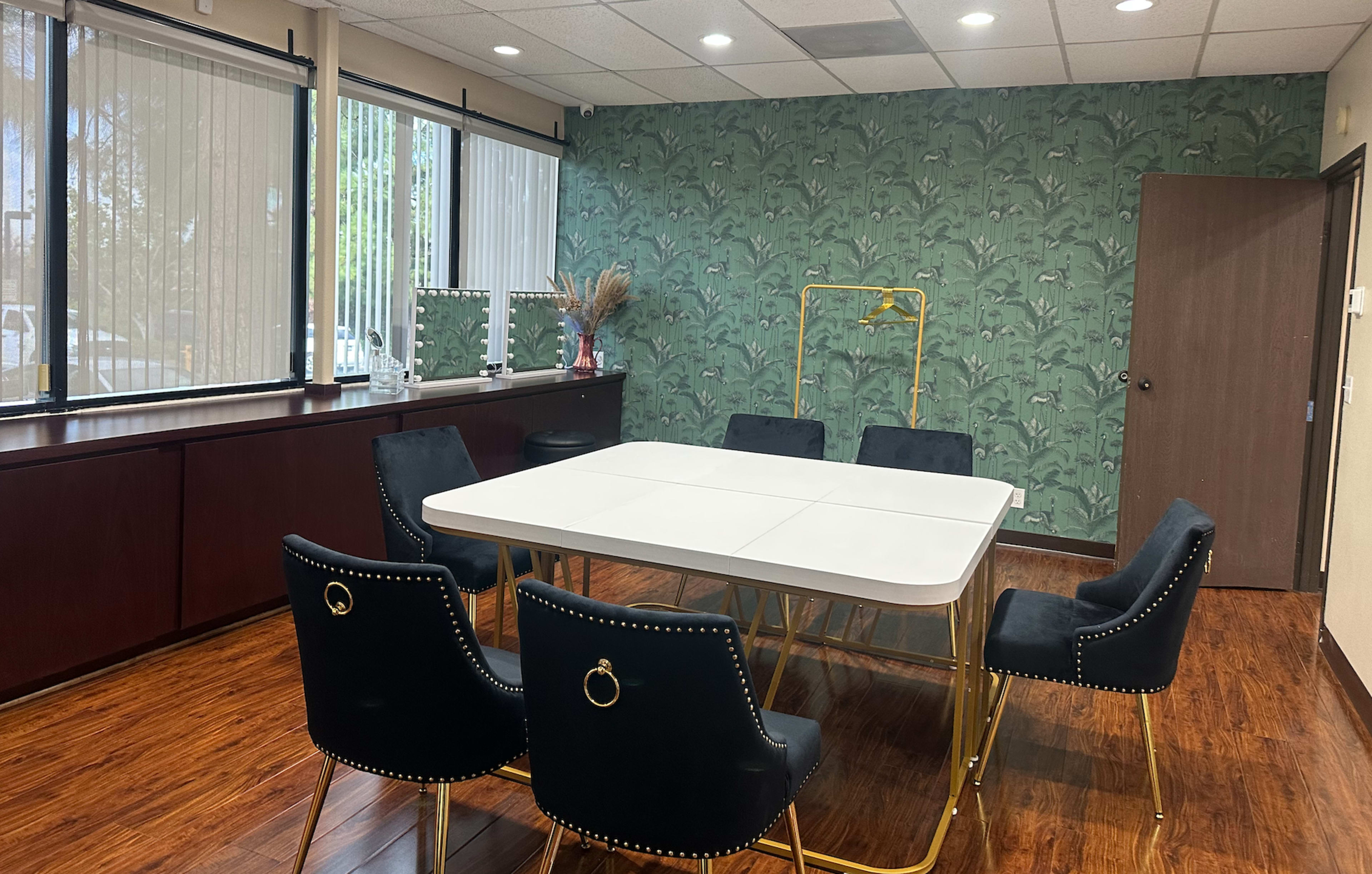 A modern meeting room featuring a white table surrounded by black chairs, with green floral wallpaper and large windows letting in natural light.