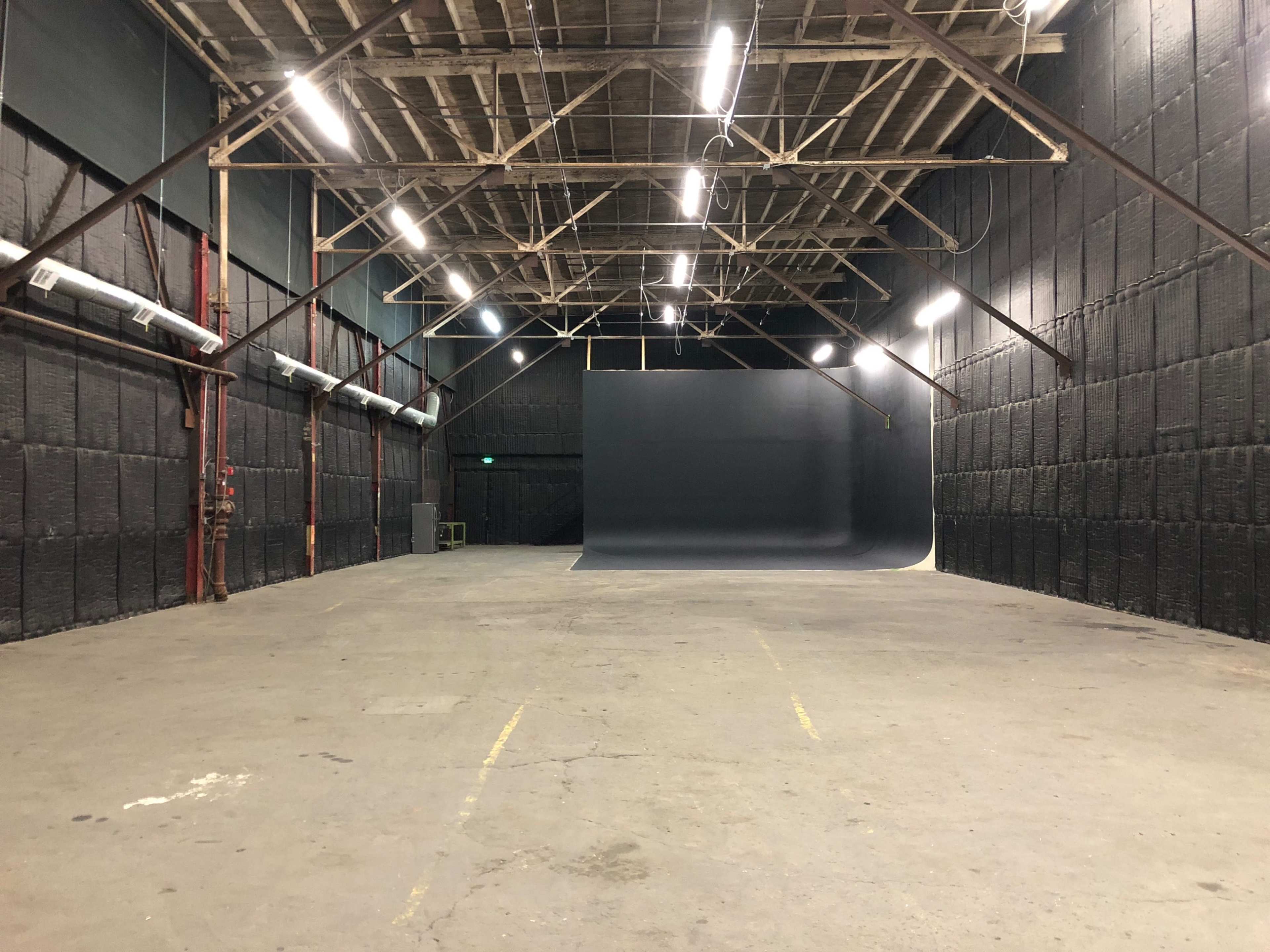 The image shows an empty studio space with high ceilings, a seamless backdrop on one end, and dim lighting.