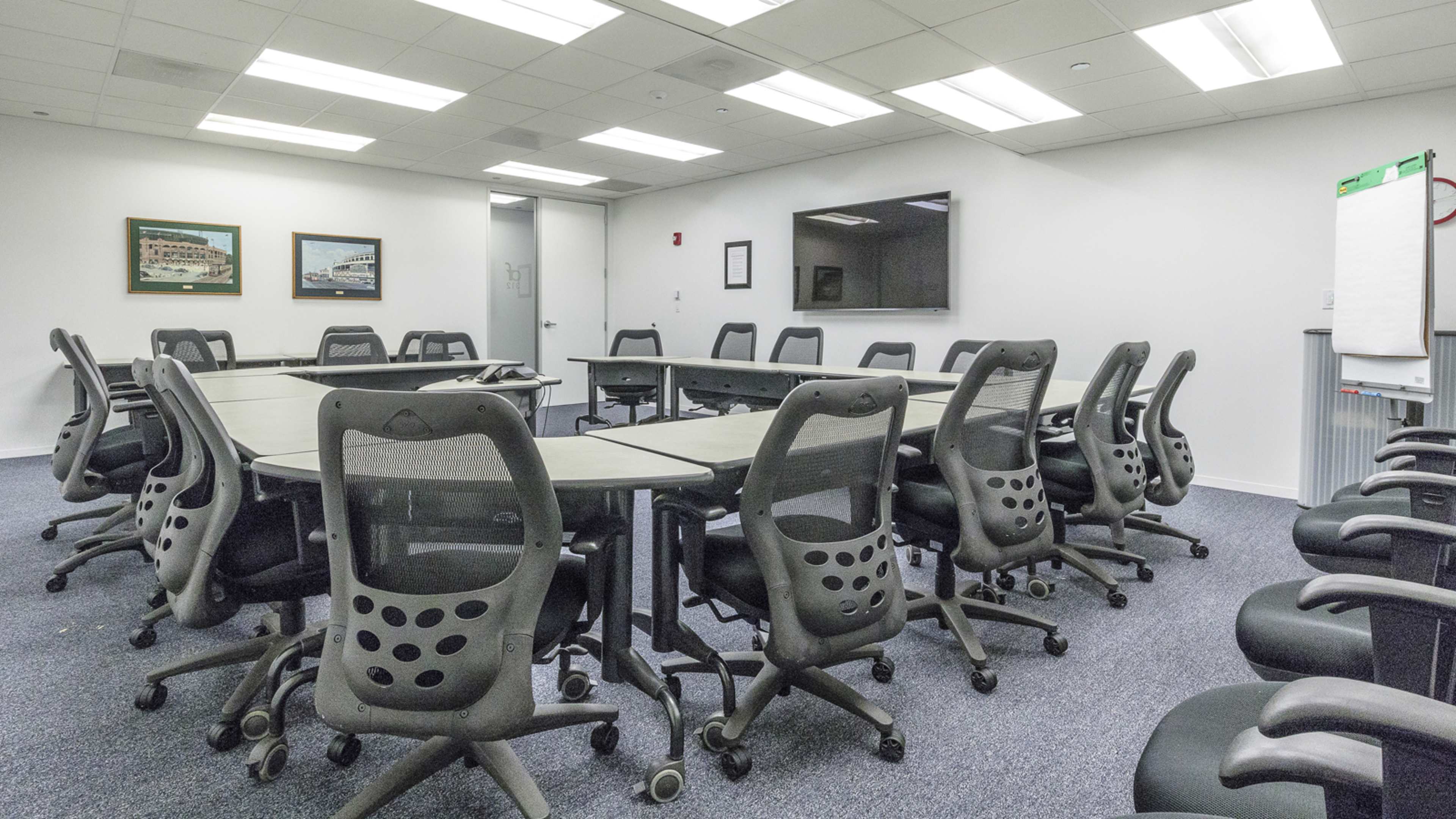 A large conference room is furnished with multiple oval tables and ergonomic black chairs arranged around them, with a TV and whiteboard on the walls.
