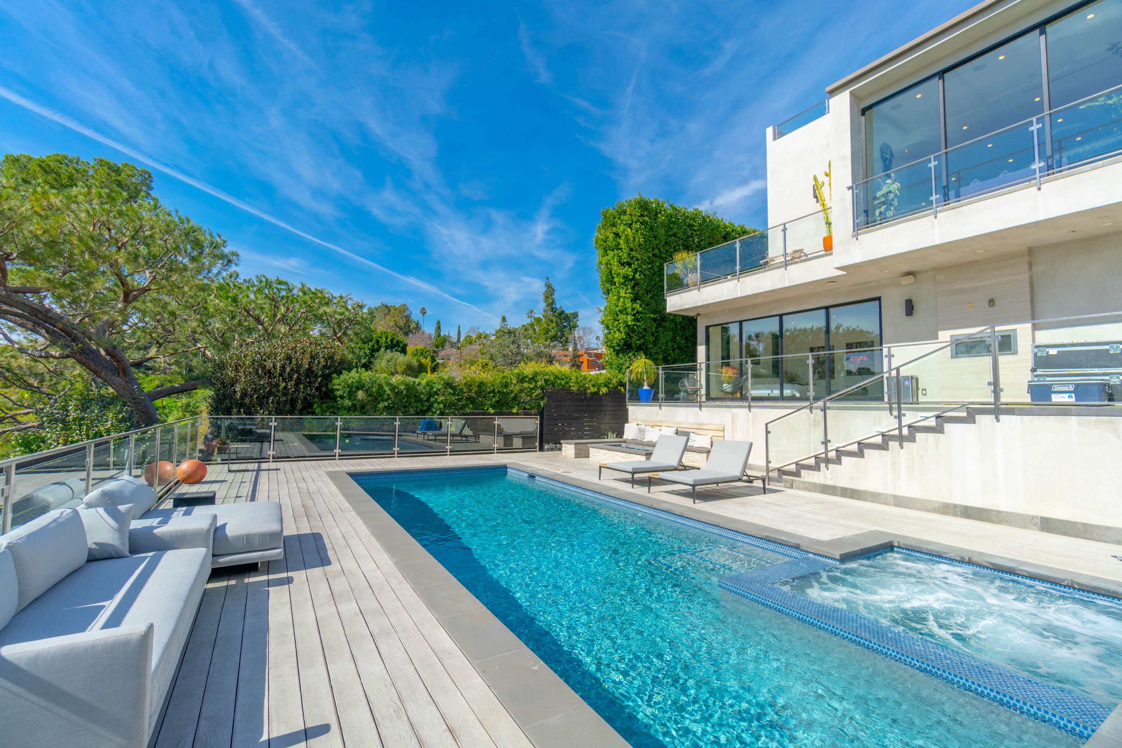 A modern house with a swimming pool and lounge area is set against a clear blue sky and lush greenery.