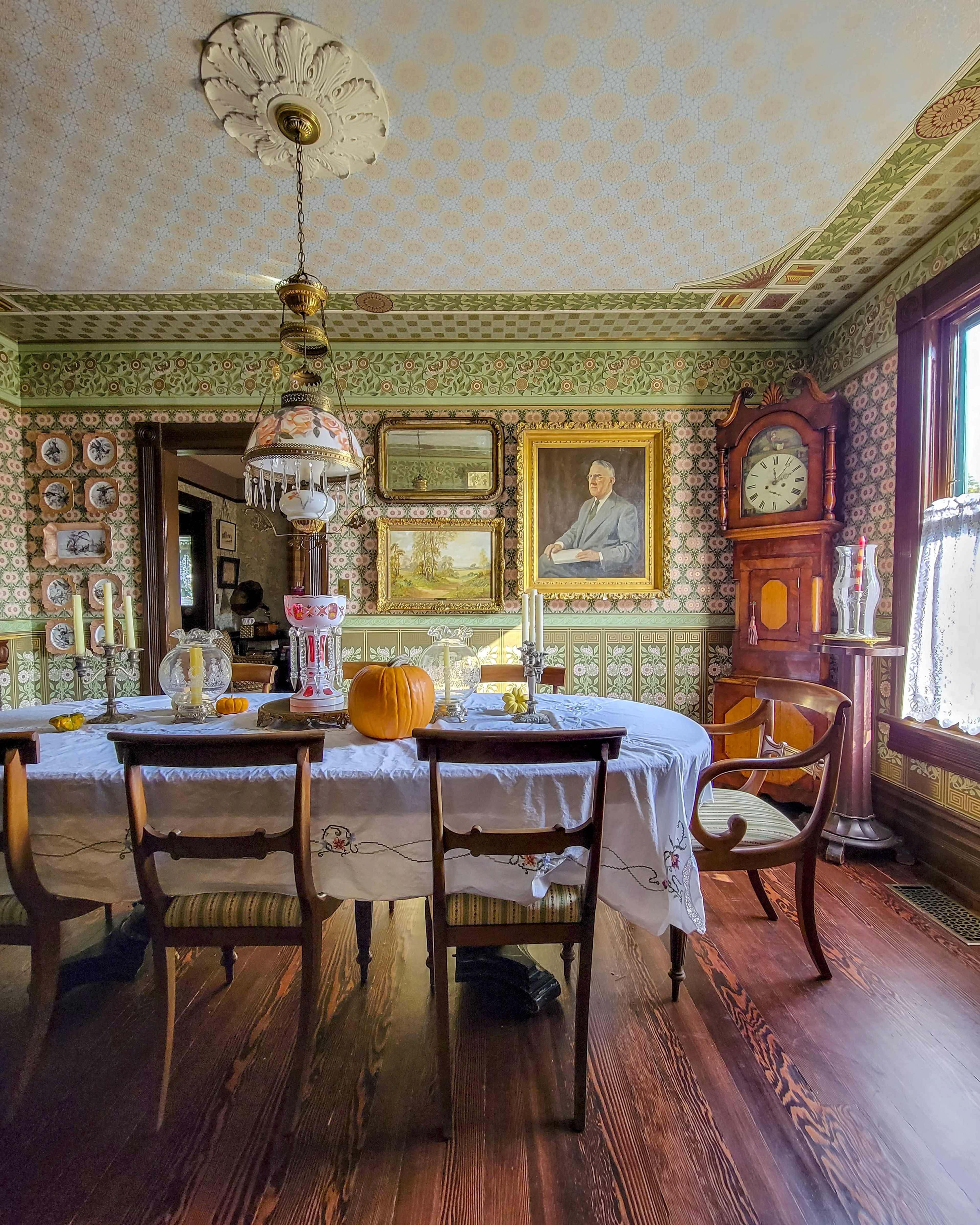 The dining room features a wooden table set with a tablecloth, surrounded by chairs, with ornate wallpaper, vintage decorations, and a large clock against the wall.