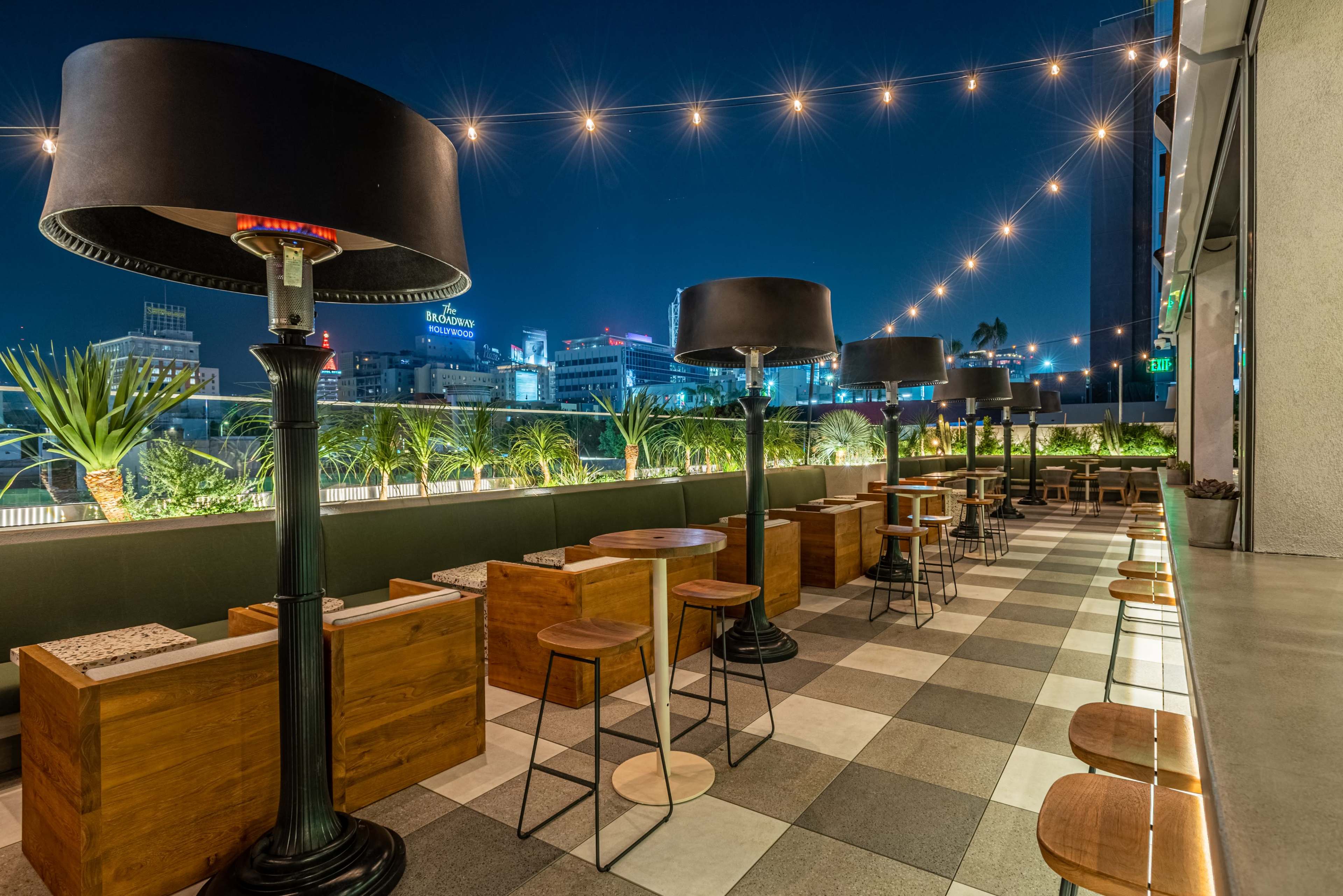 The image shows a modern outdoor bar area with wooden seating, large lamps, and decorative plants under string lights in a cityscape at night.