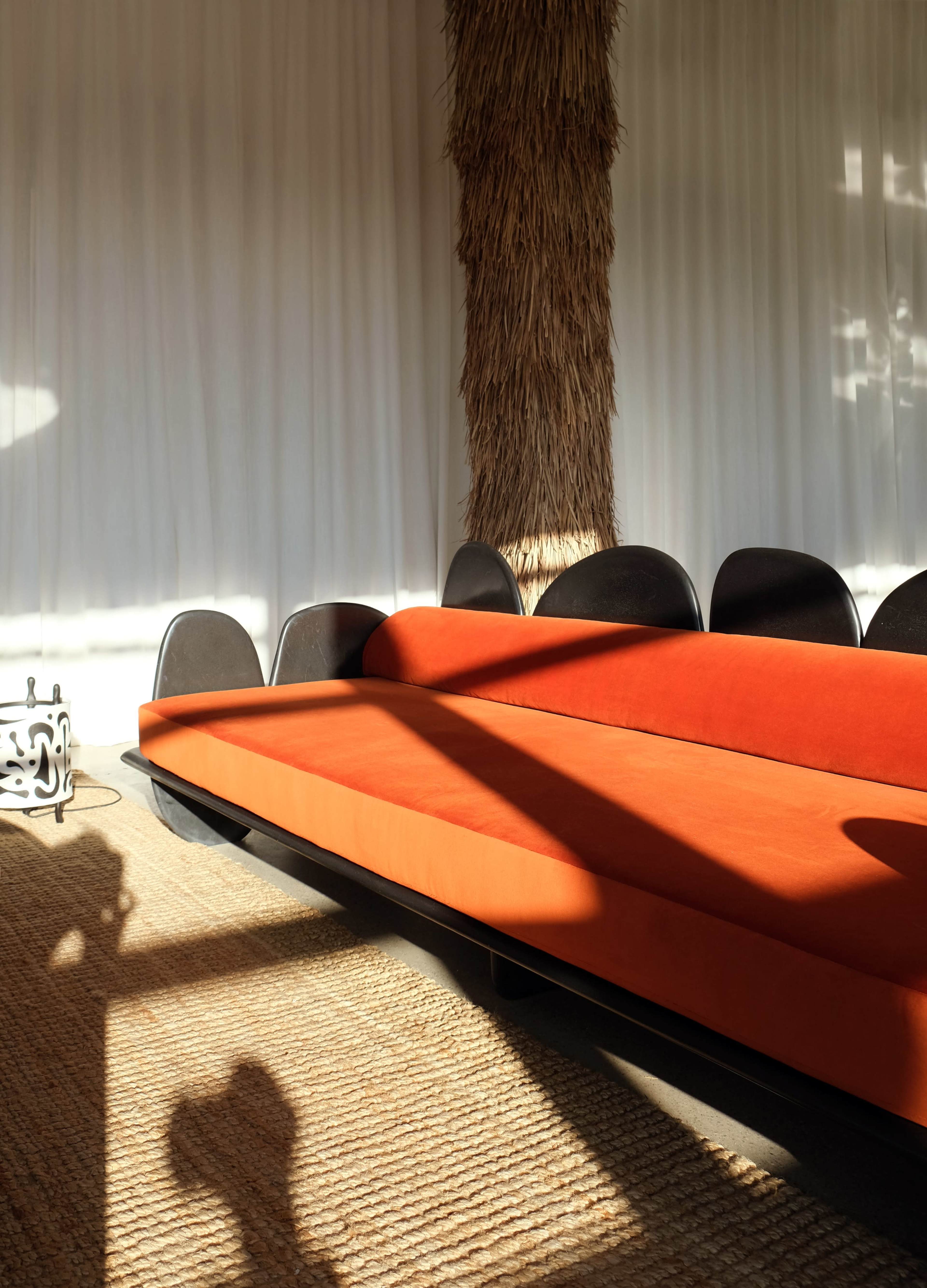 The image shows a modern orange sofa positioned against a wall with white curtains, accompanied by black chairs and a textured area rug on the floor.