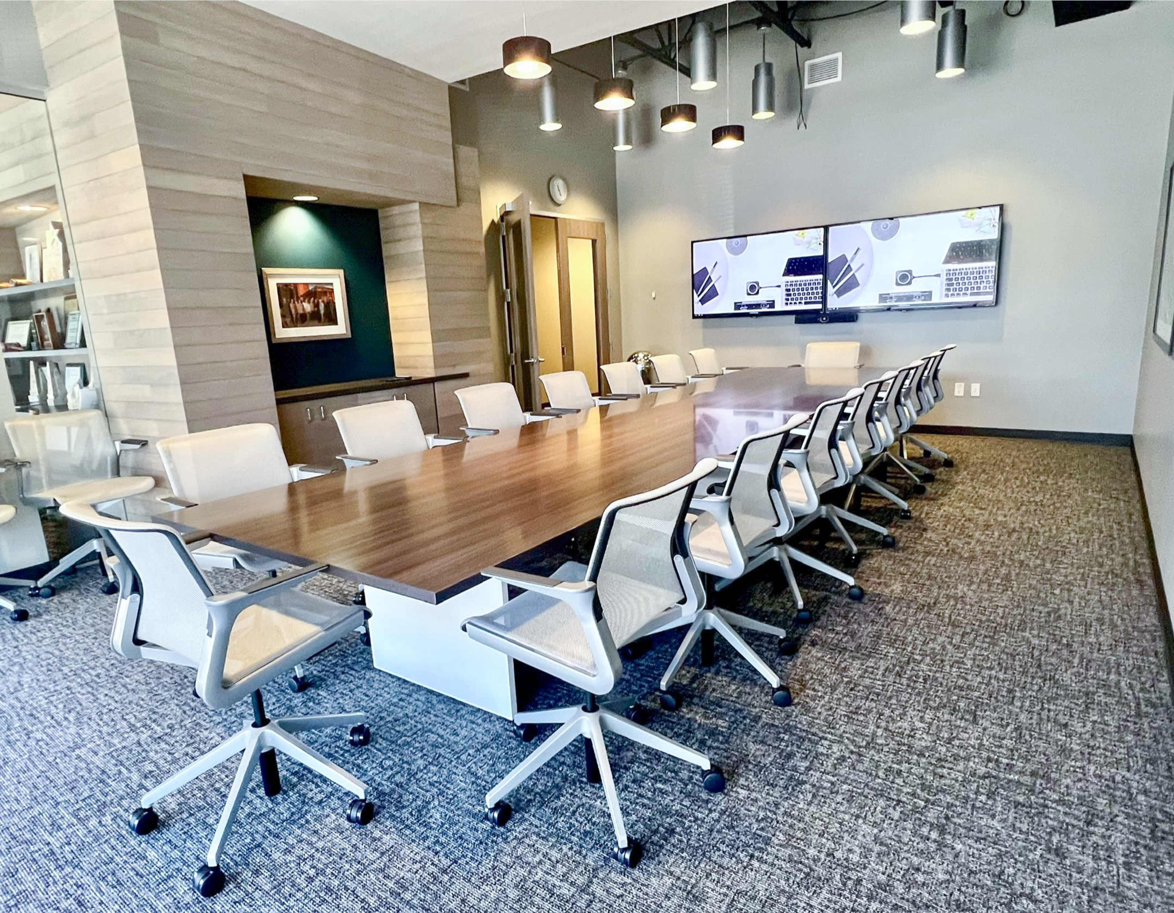 A modern conference room features a long wooden table surrounded by ergonomic chairs and two large screens displaying computers on a gray wall.